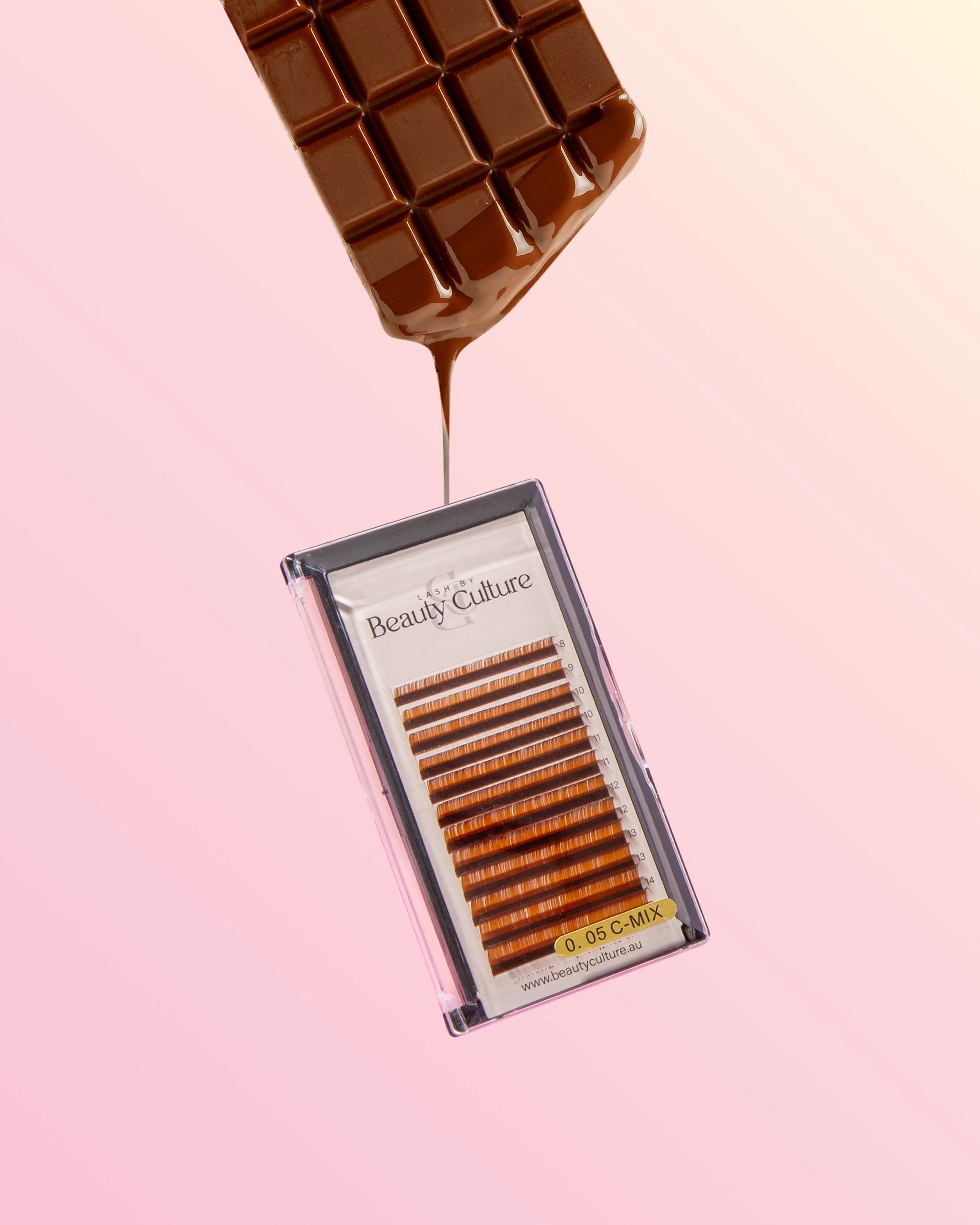 A bar of milk chocolate melting with chocolate dripping down onto a box of eyelash extensions labeled 'Beauty Culture' on a pink background.