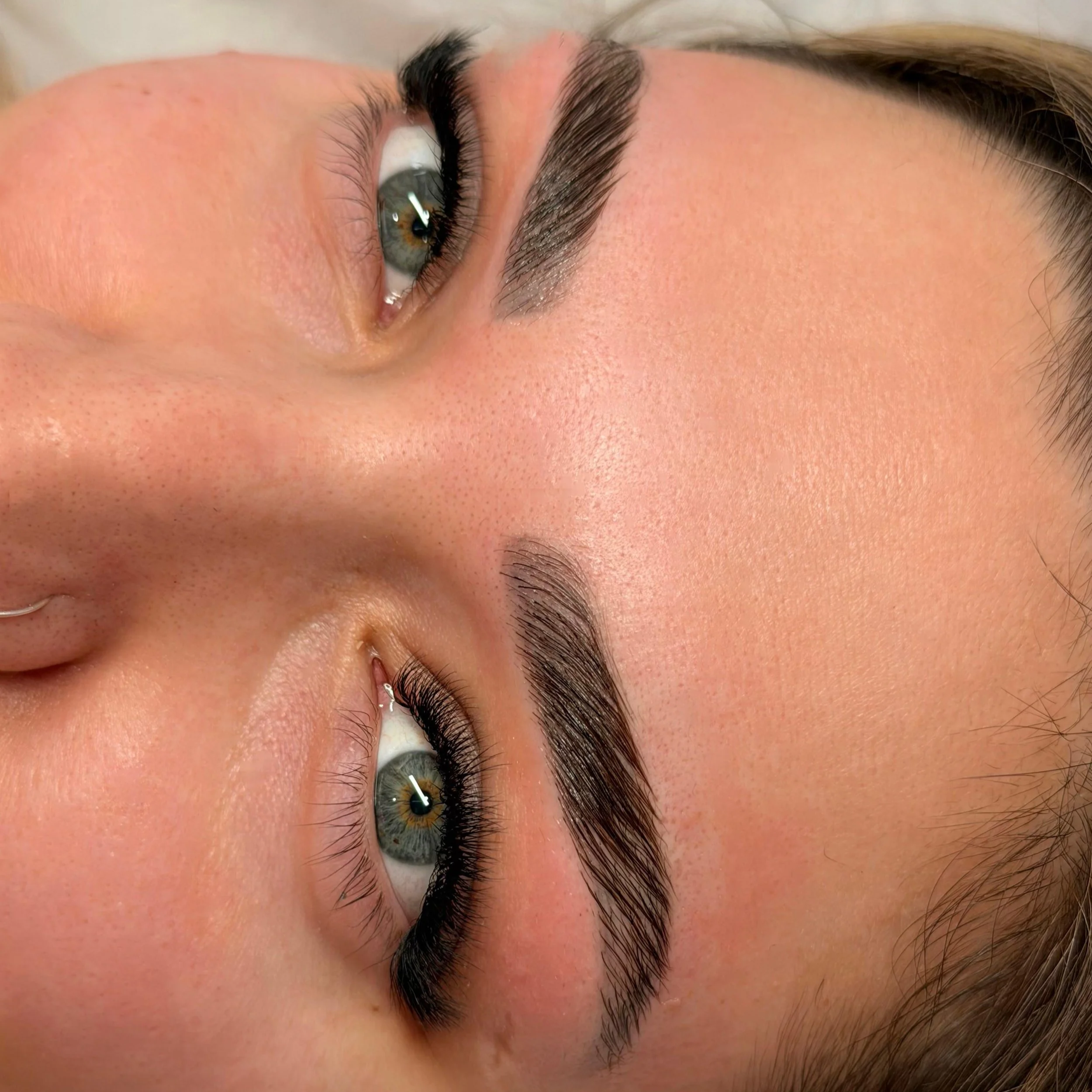 Close-up of a woman's face with well-groomed eyebrows, light skin, and blue eyes, lying down with her eyes looking upward.