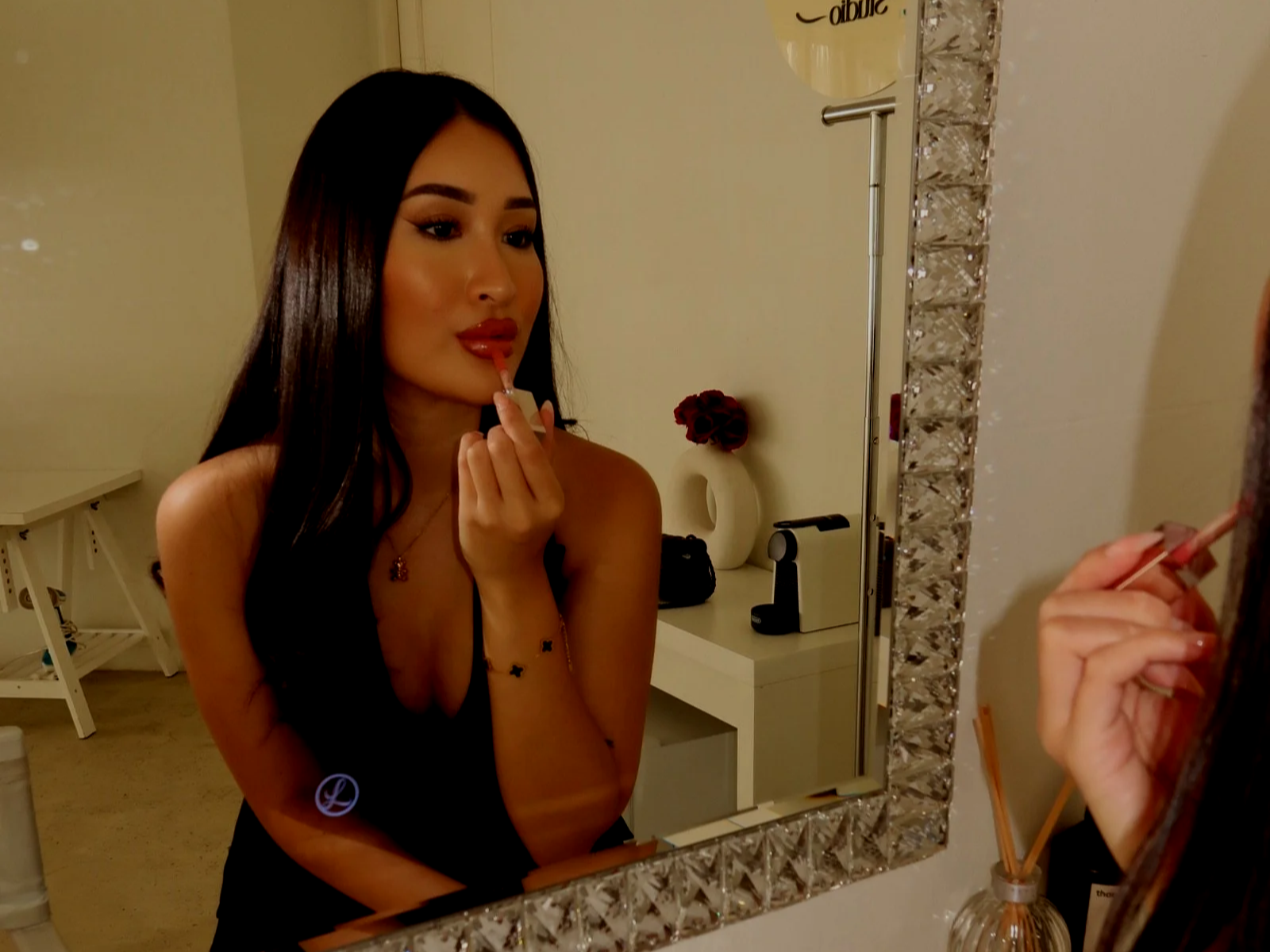 A woman with long dark hair applying lipstick while looking into a decorative mirror in a room with a white dresser, a vase with red roses, and various objects.