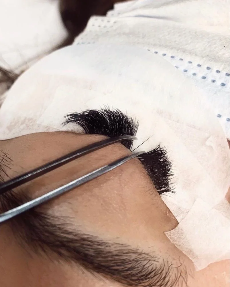 Close-up of eyelash extension procedure with tools on a person's eye, with tissue paper nearby.