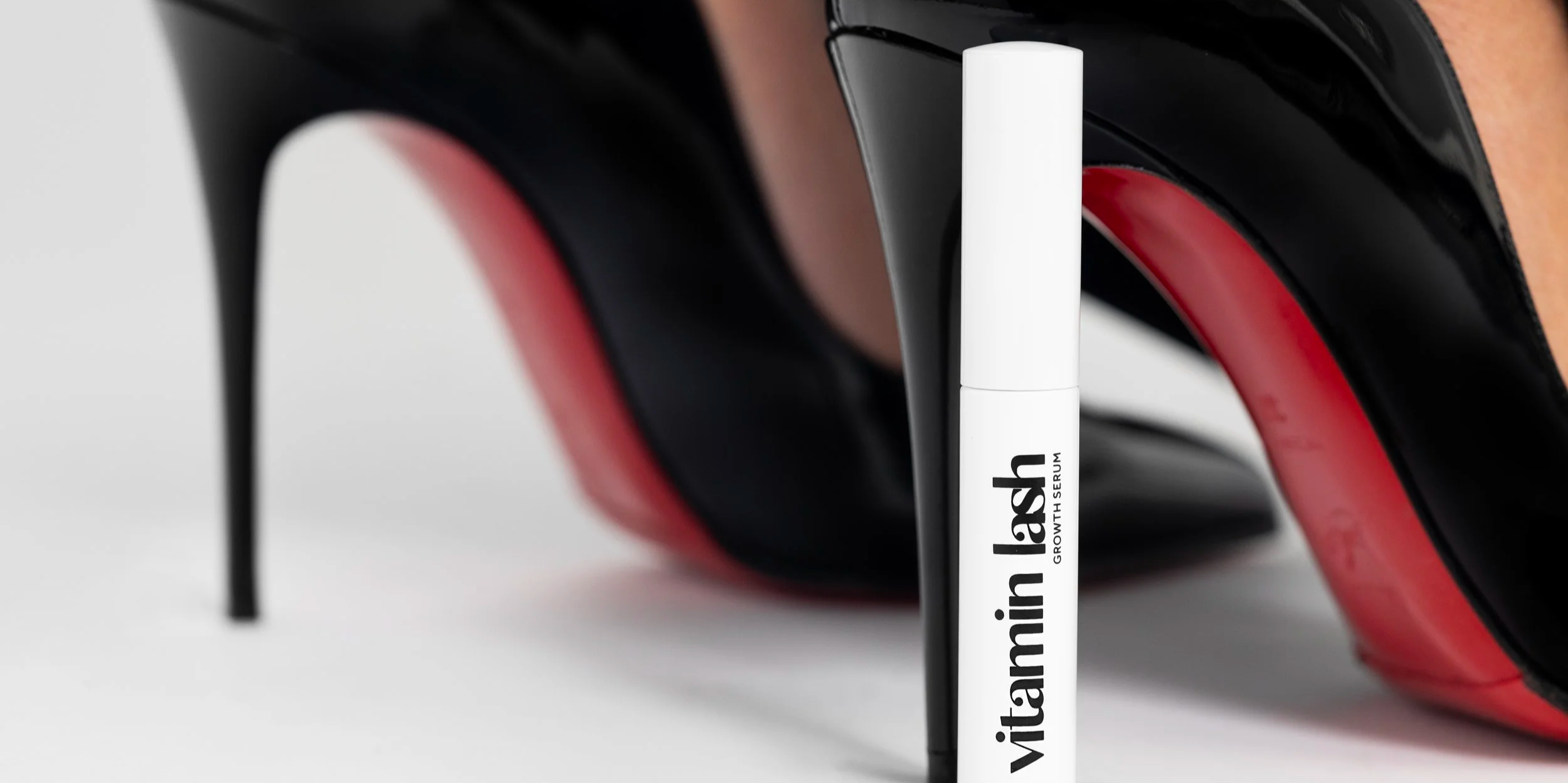 Close-up of a white felt-tip makeup eyeliner pen with black text labeling 'vitamin lash growth serum' in front of a pair of black high-heeled shoes with red soles.