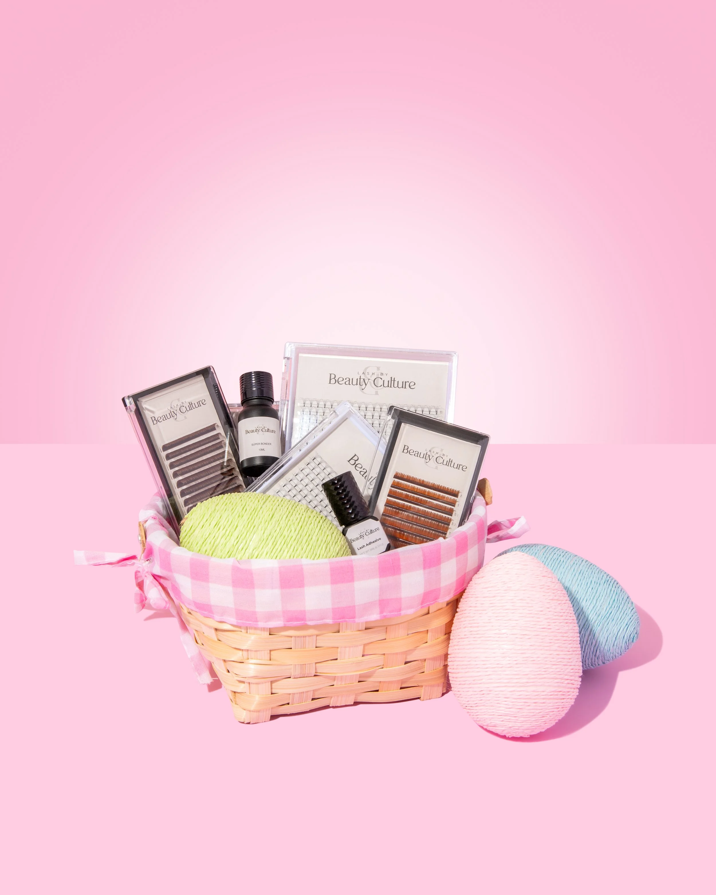 Eyelash extension kit with various supplies in a pink checkered basket and three decorative eggs on a pink background.