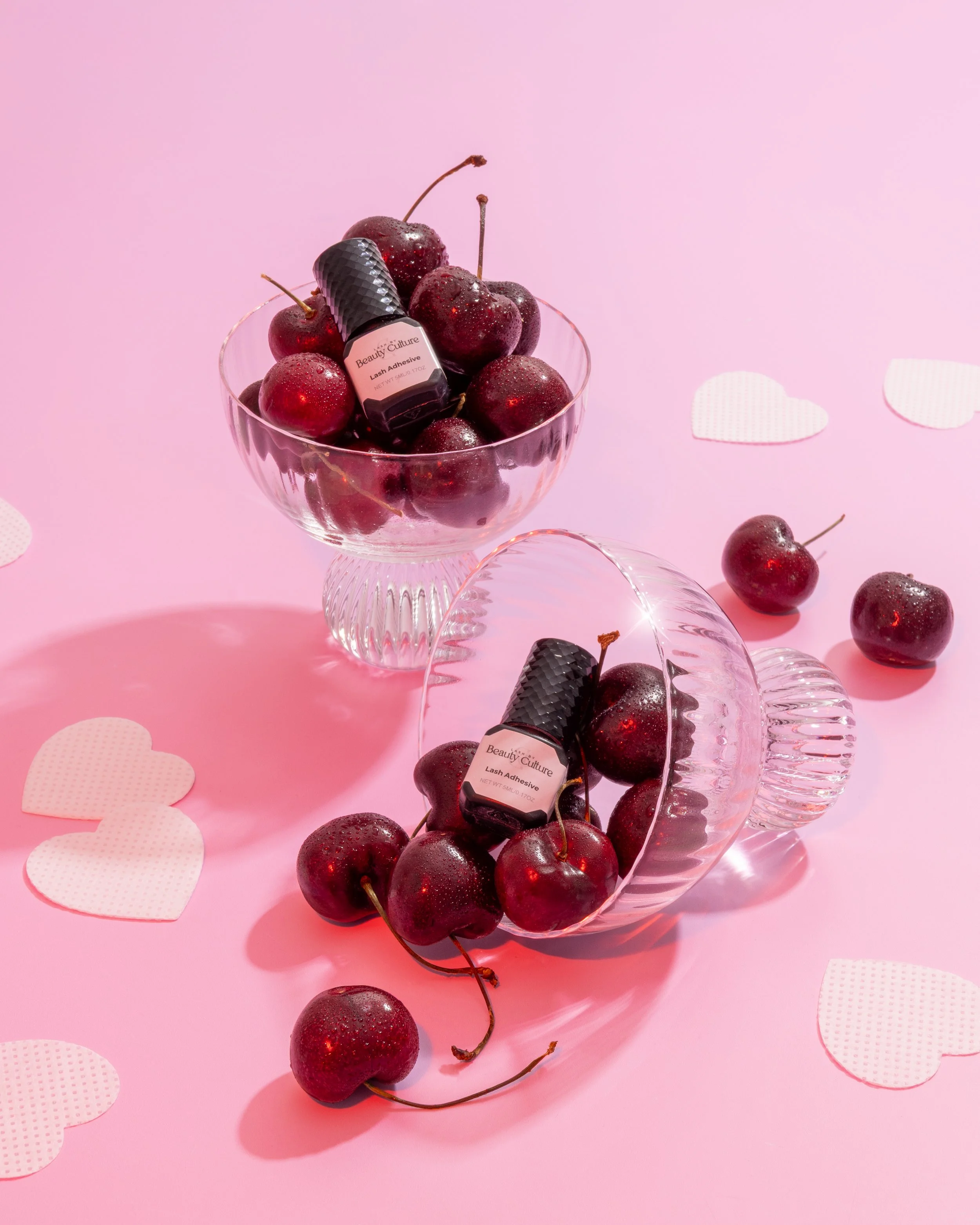 Two glass bowls filled with cherries, with a small bottle of eyelash adhesive placed on top of the cherries in each bowl, set on a pink background with paper heart decorations.