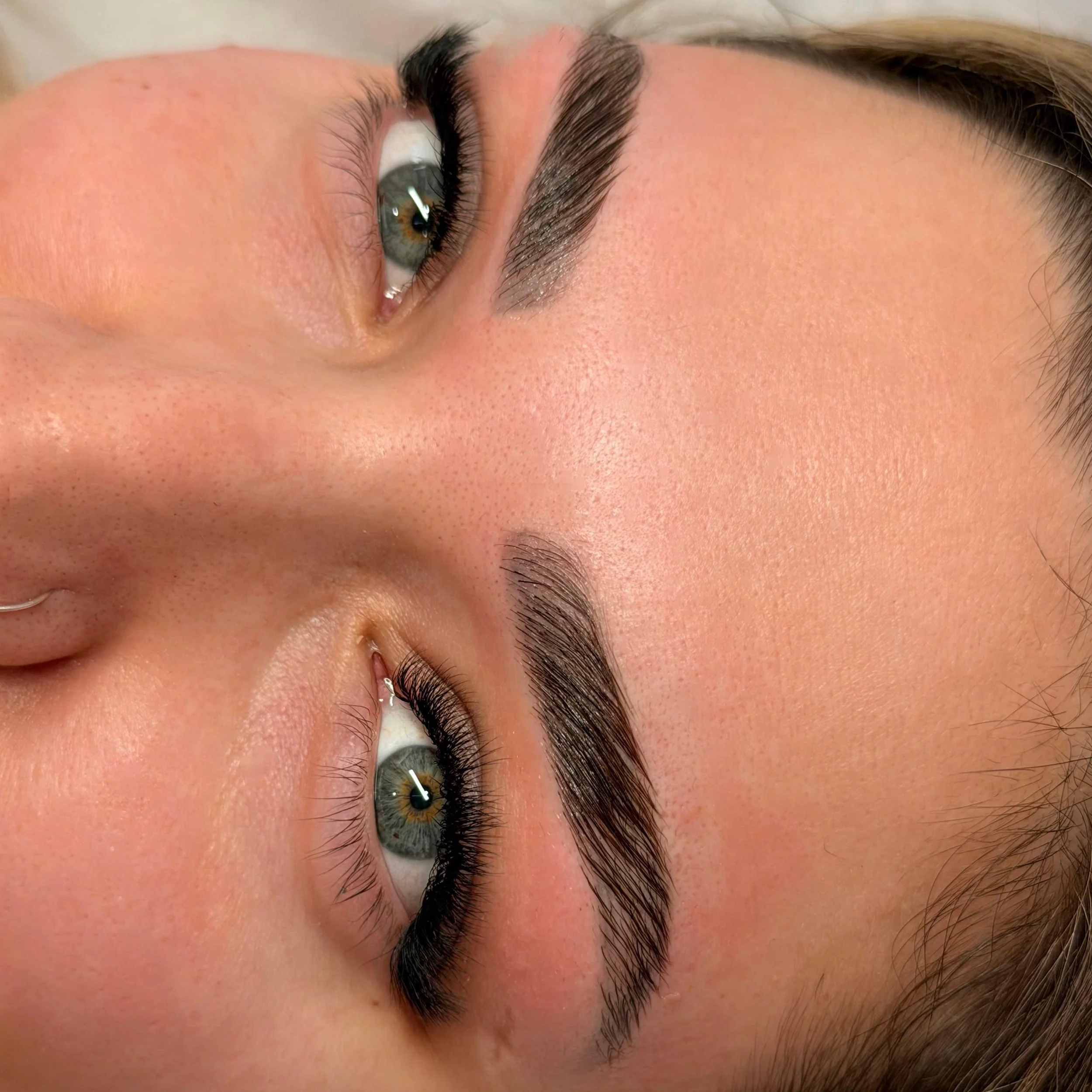 Close-up of a person's face lying down, showing well-groomed eyebrows, eye makeup with eyeliner, and hazel eyes.