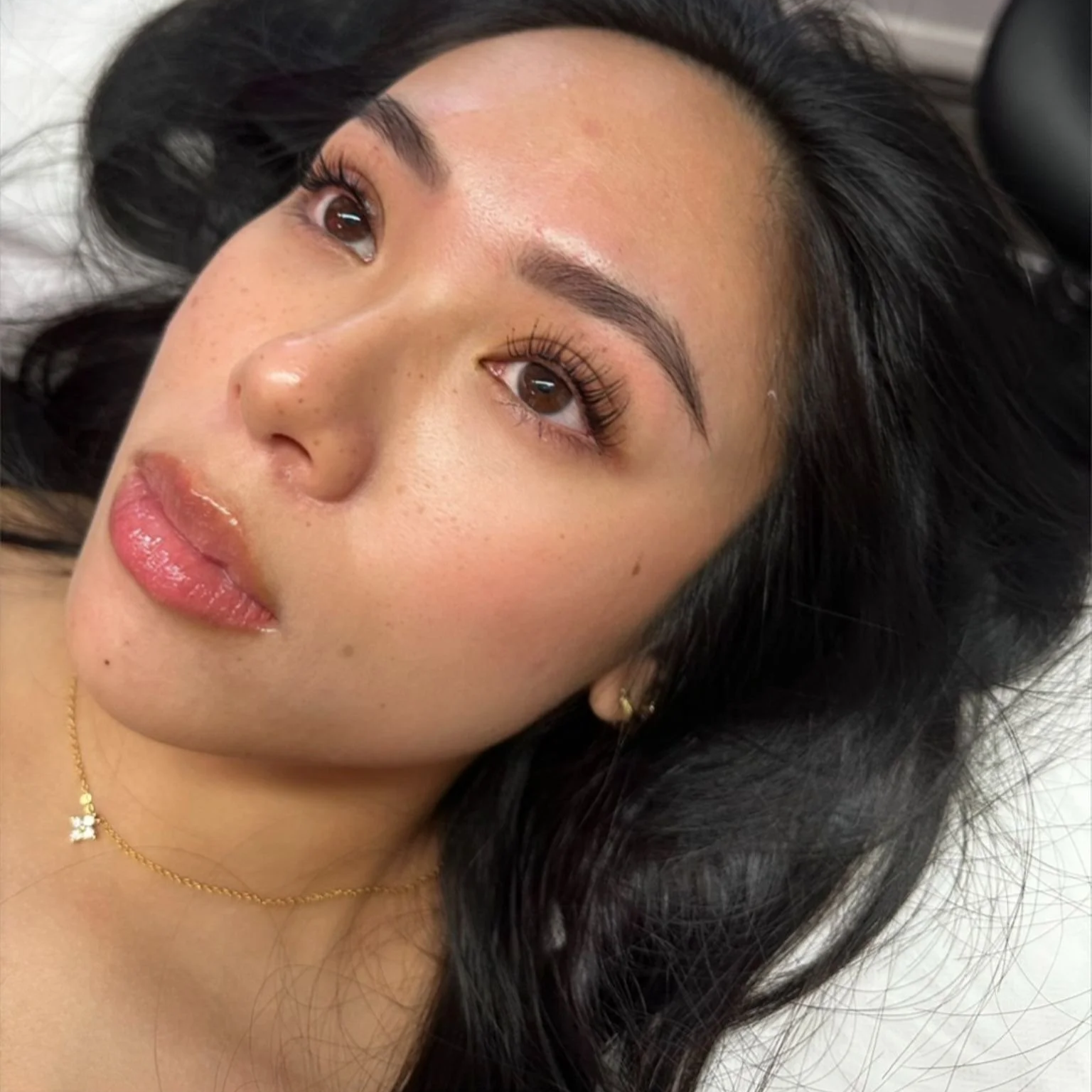 Close-up of a young woman with dark hair lying down, showing her face with makeup, earrings, and a gold necklace.