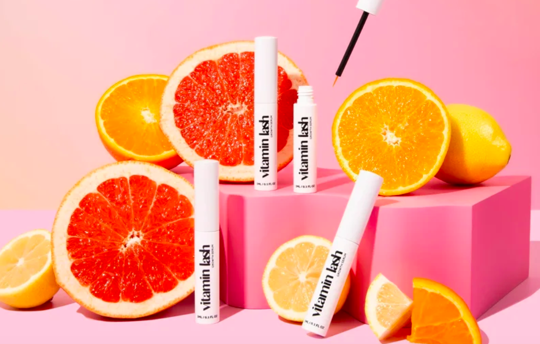 Cosmetics tube labeled 'Vitamin Lash' surrounded by sliced grapefruit and lemon on pink background.