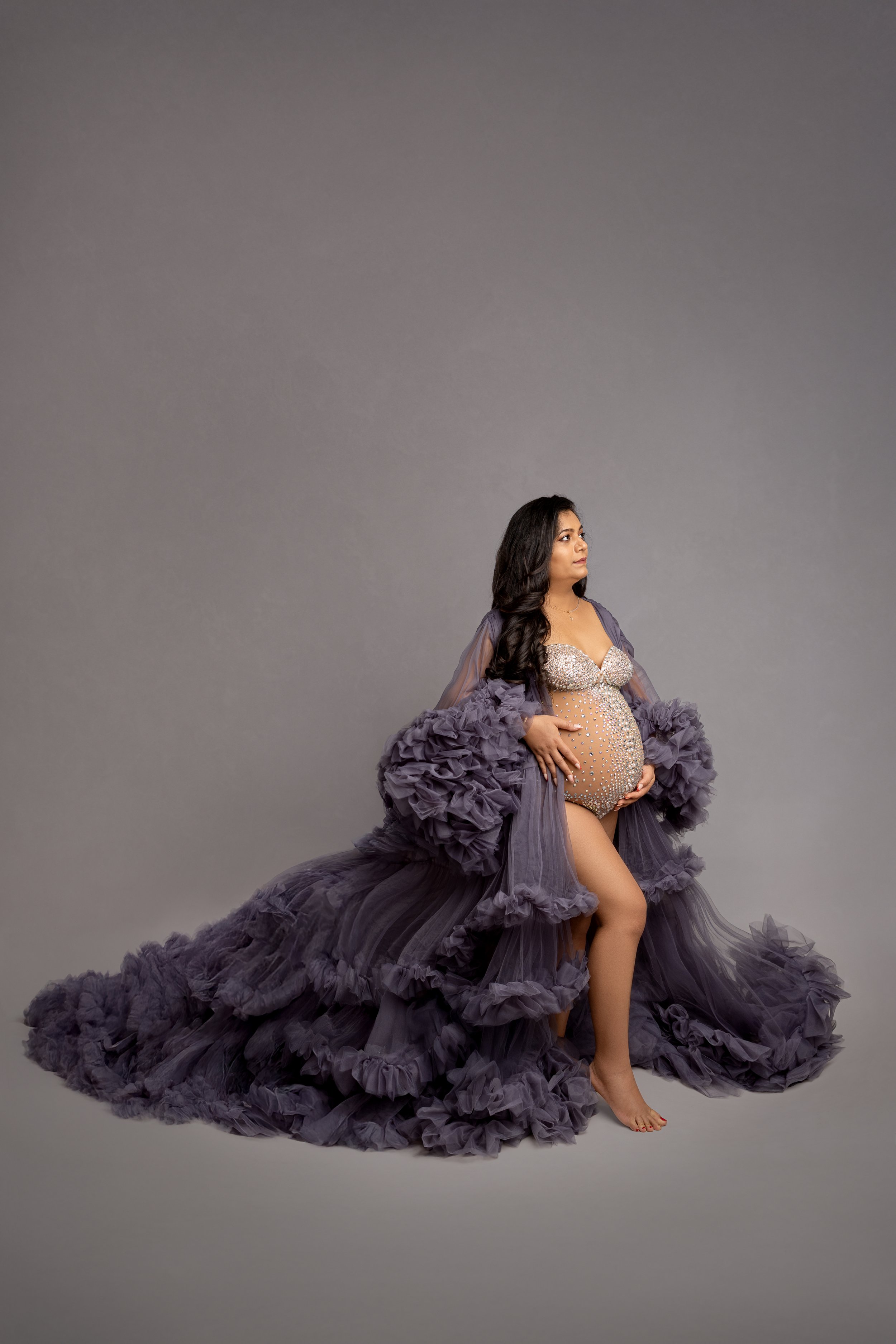 Pregnant woman in an elaborate purple gown with ruffles and tulle, sitting against a plain gray background.