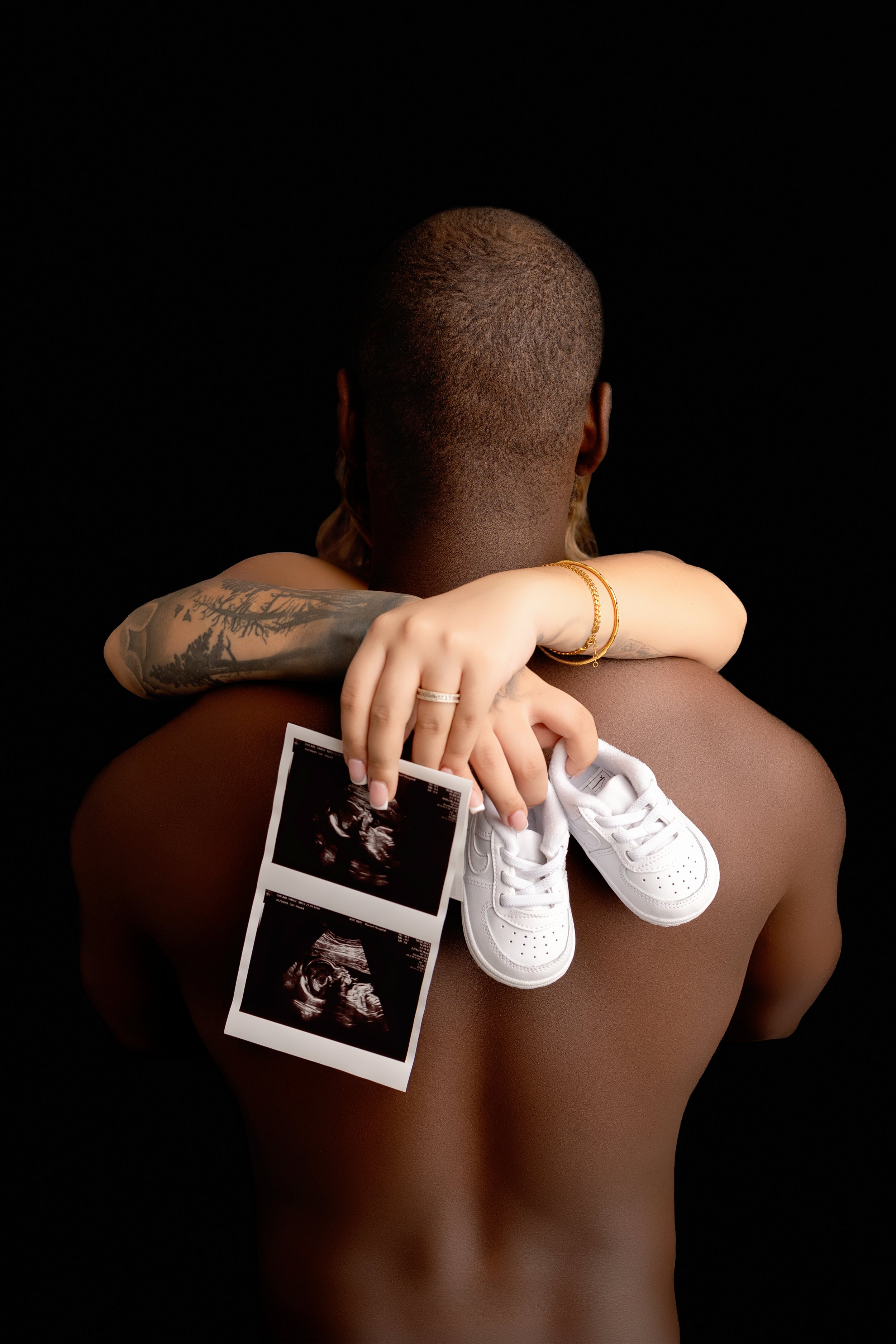 A shirtless person with a shaved head holding a small baby in their arms. The person is holding ultrasound images and a pair of tiny white baby shoes.