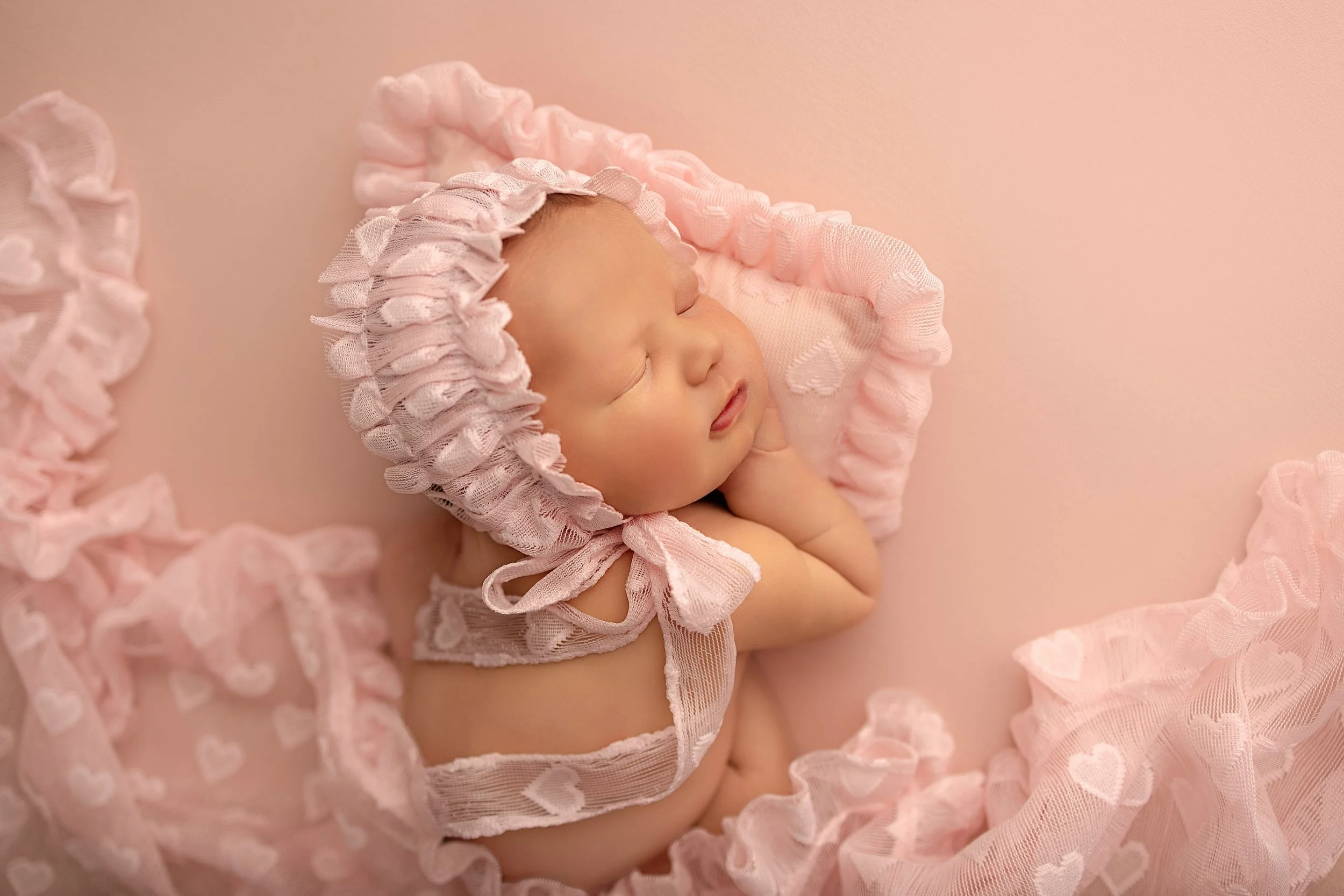 A sleeping newborn baby wearing a pink bonnet with ruffles and a matching pink blanket with heart patterns, lying on a soft pink surface, with one hand tucked under the chin.