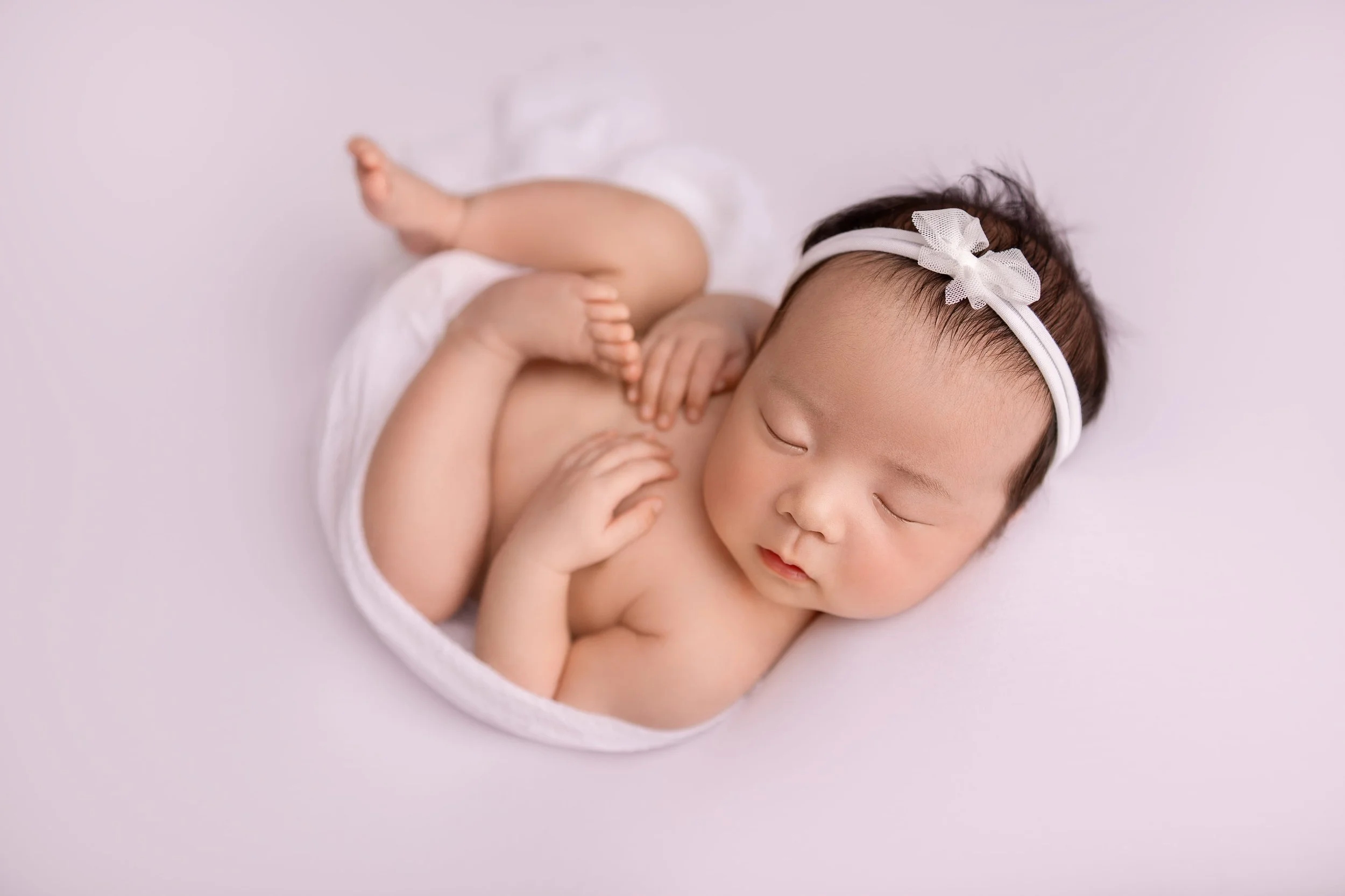 Sleeping baby girl with a white headband decorated with a bow, lying on a soft pink surface, partially covered with a white cloth.