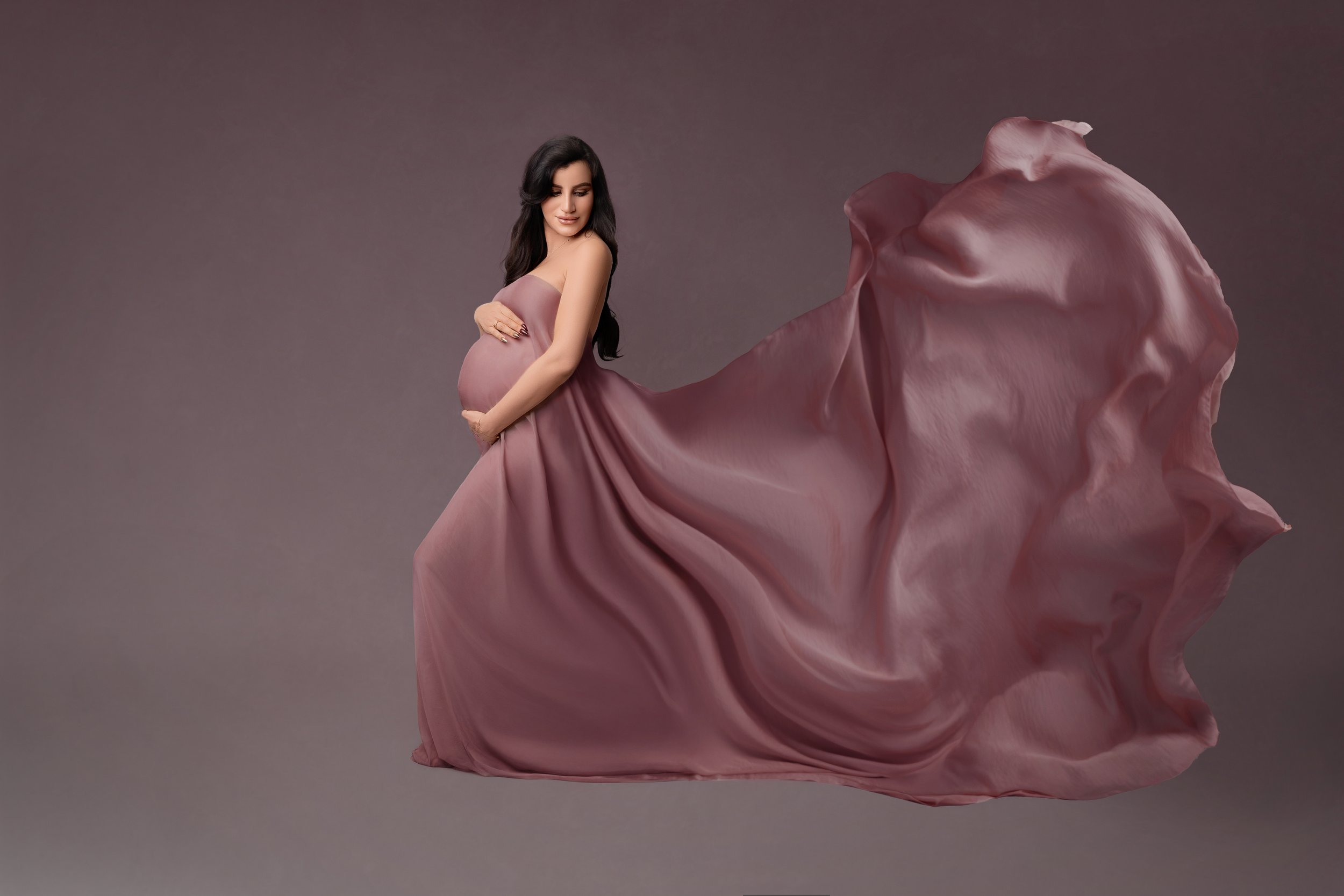Pregnant woman in a flowing mauve gown standing with hands on her belly against a neutral background.