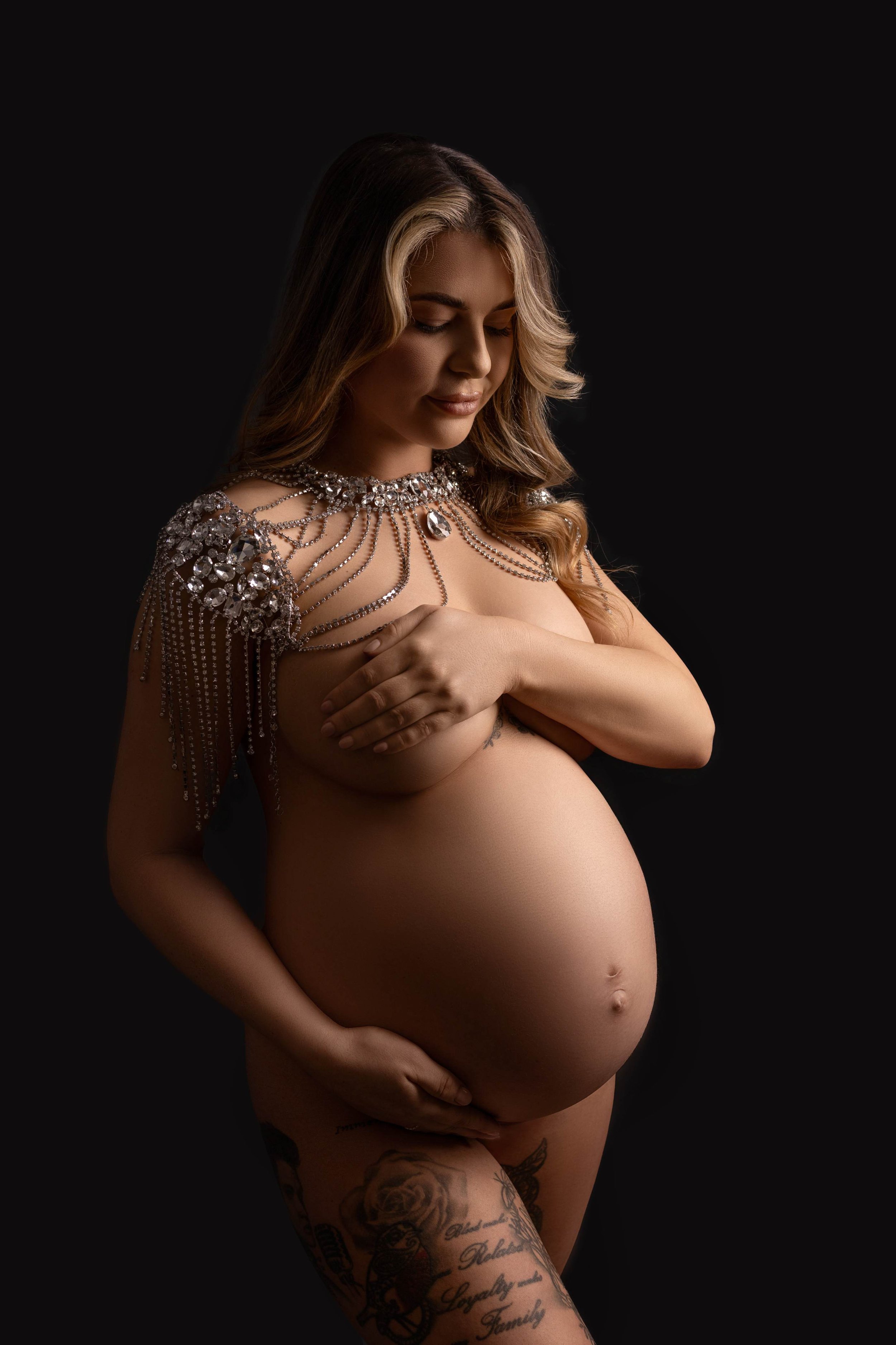 A pregnant woman with long, wavy blonde hair poses against a black background. She is topless, covering her chest with one hand, and wearing an ornate, bejeweled shoulder covering. She has tattoos on her thigh and lower abdomen.