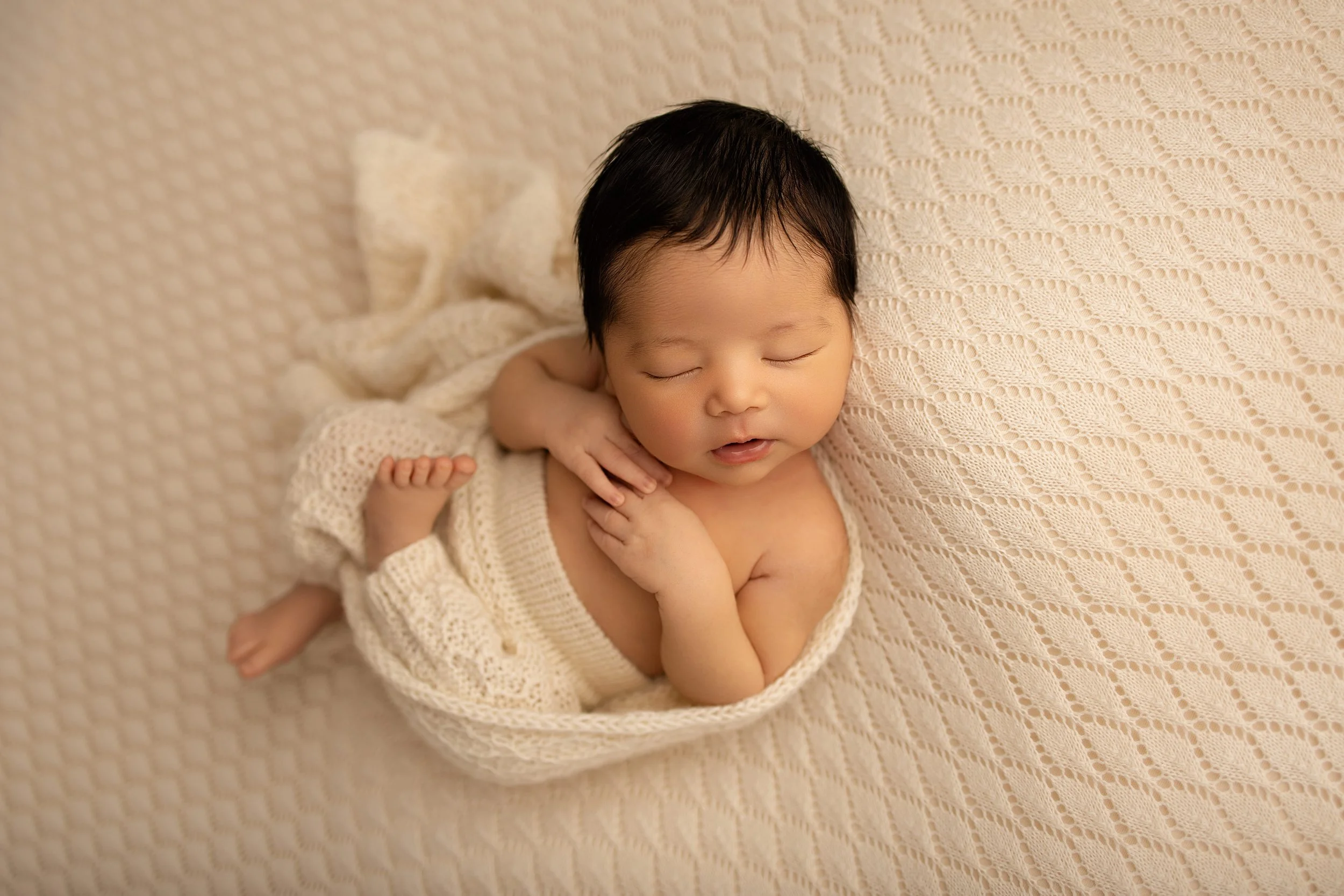 A sleeping baby with black hair, resting on a textured beige blanket, partially wrapped in a knitted cream-colored blanket.