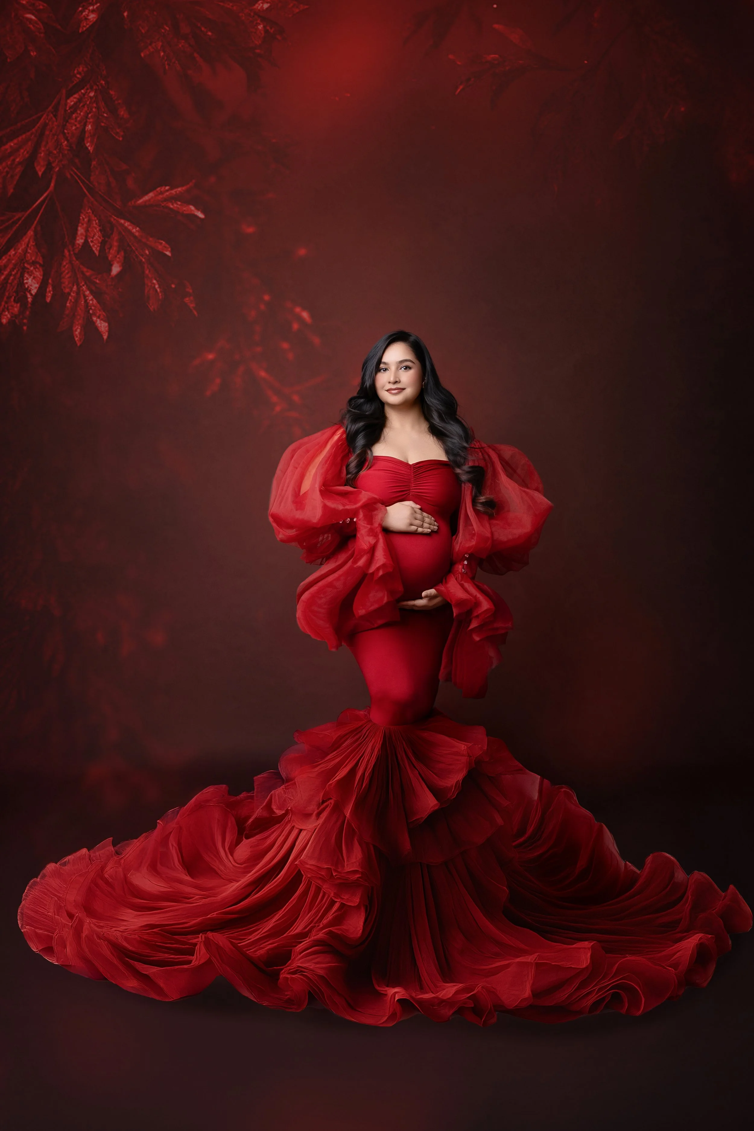 A pregnant woman wearing a red, ruffled, flowing gown standing in front of a red, leafy background.