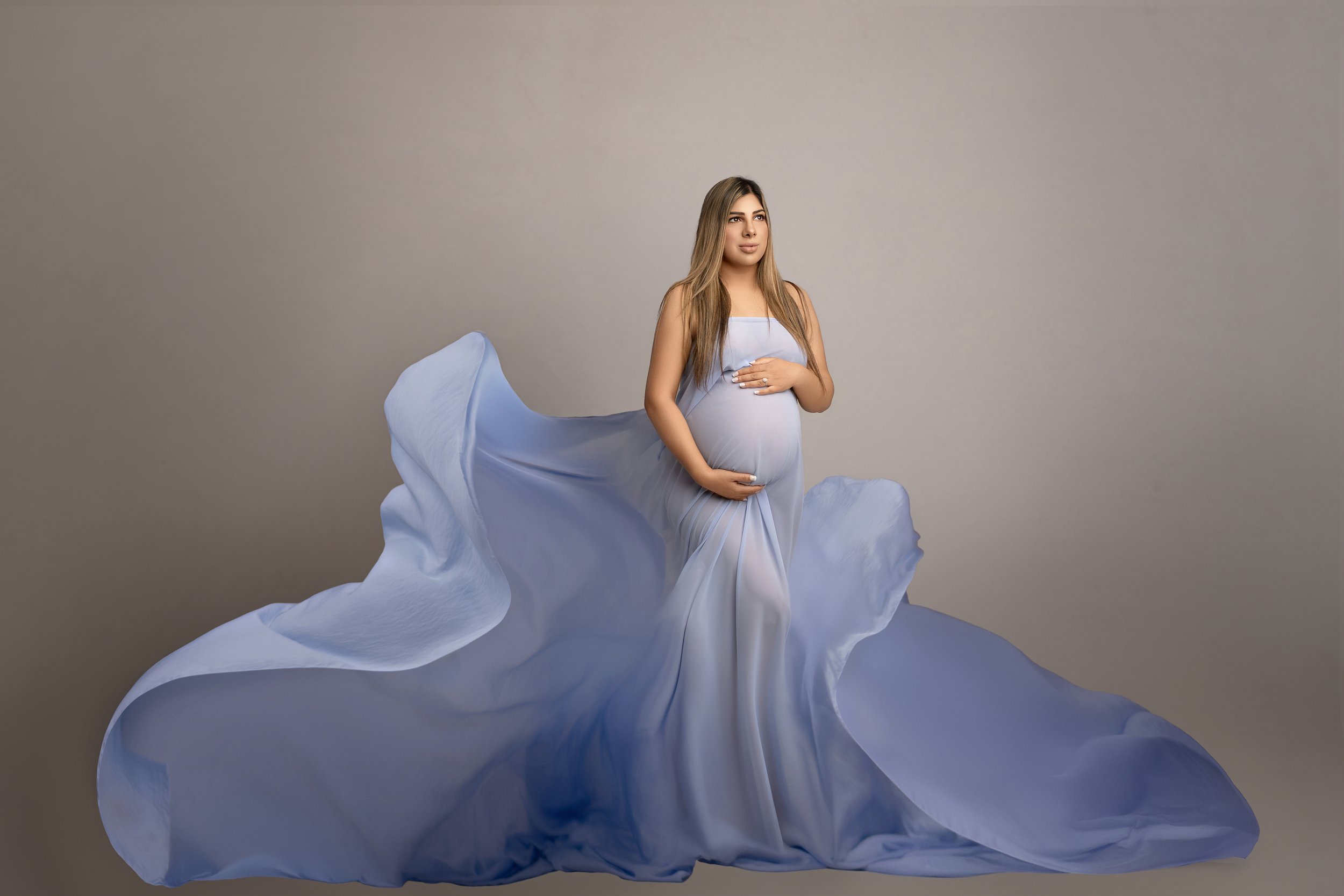 A pregnant woman in a flowing, light blue dress standing against a neutral background, with her hands gently on her belly and her gaze turned slightly to the side.