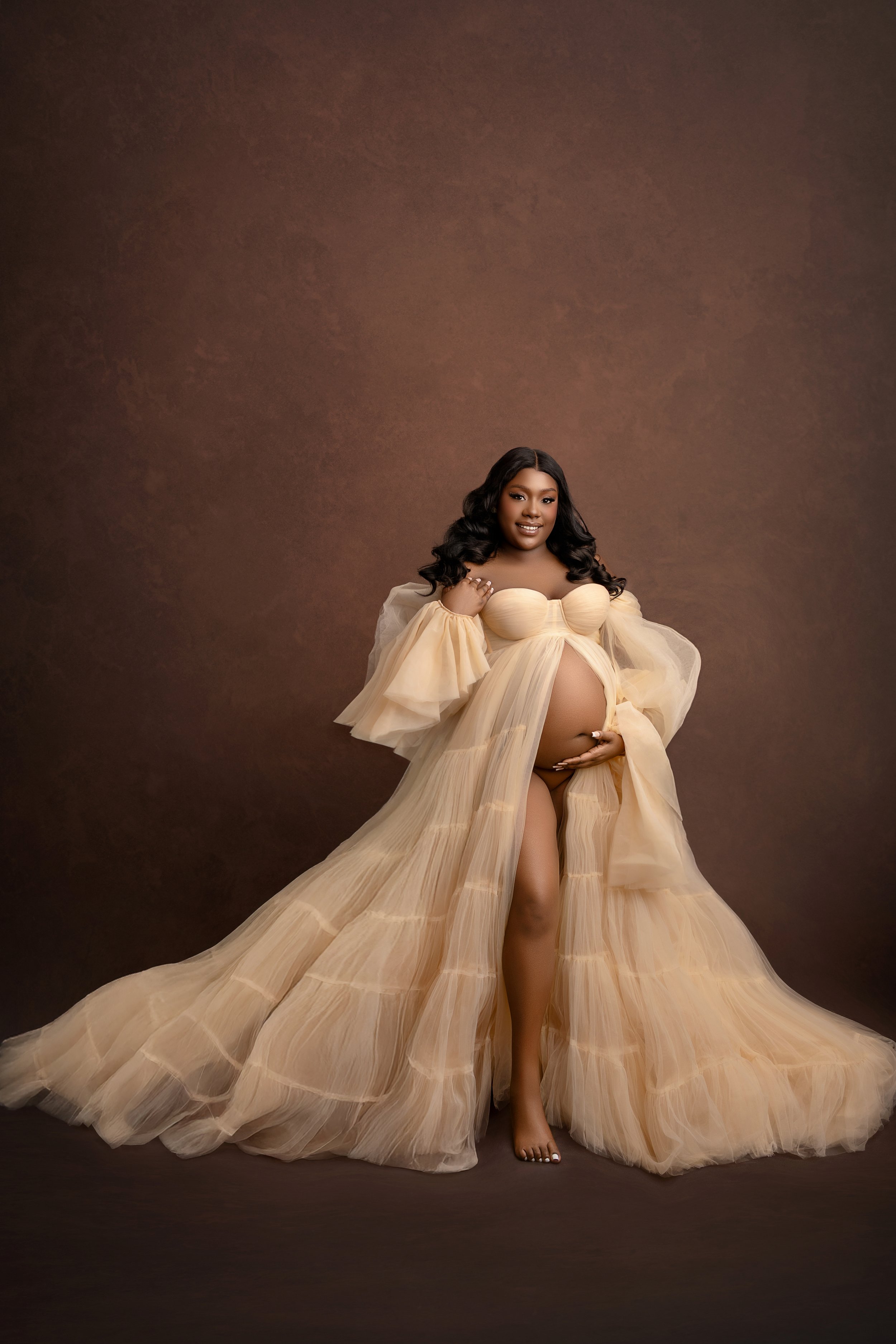 Pregnant woman in a flowing beige gown with a high slit, standing against a brown backdrop, smiling at the camera.
