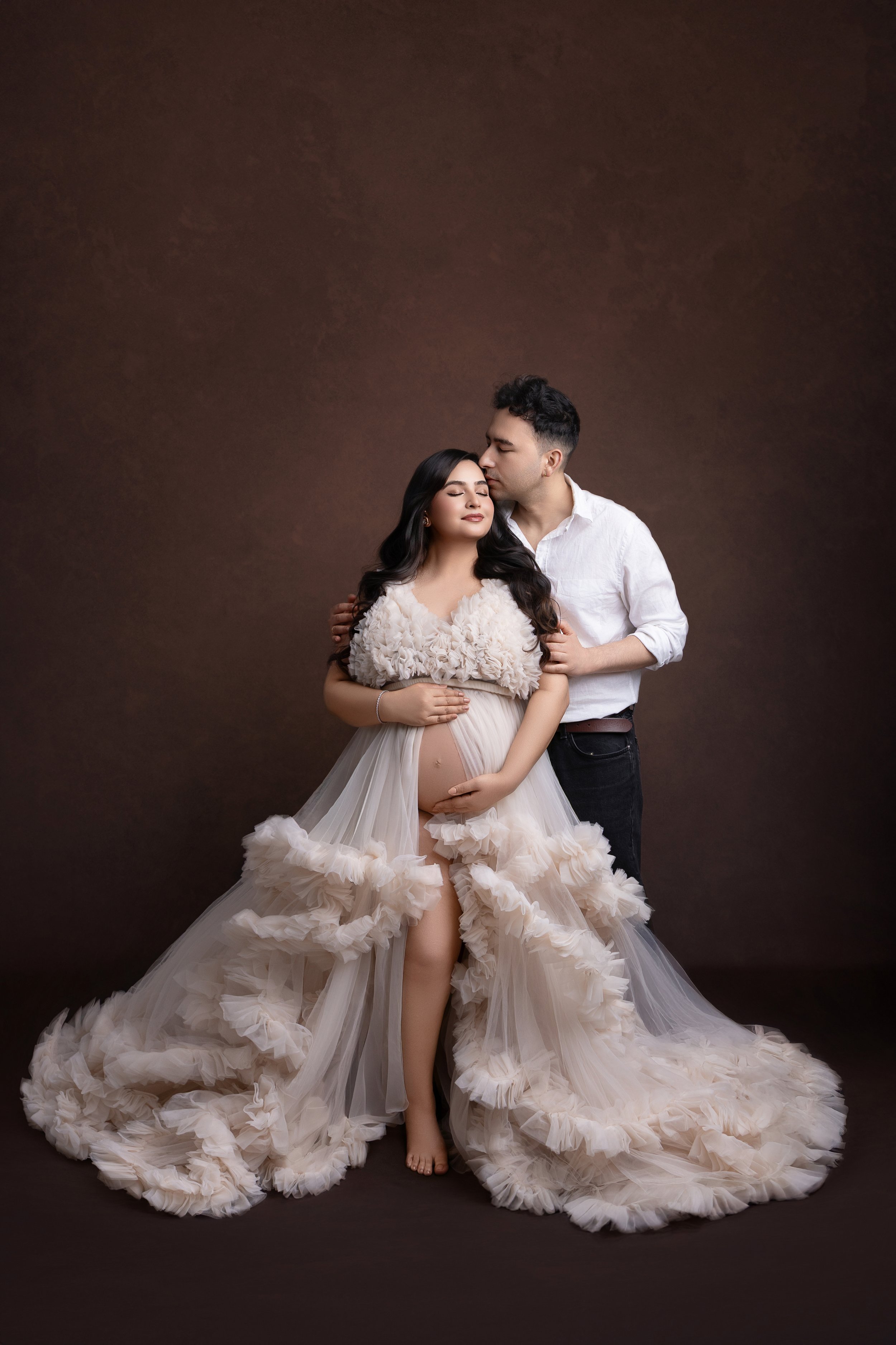 A pregnant woman in a flowing, ruffled dress poses with her partner against a dark brown background. The woman has long dark hair and appears serene with closed eyes, while the man, dressed in a white shirt and dark pants, gently kisses her head, emb
