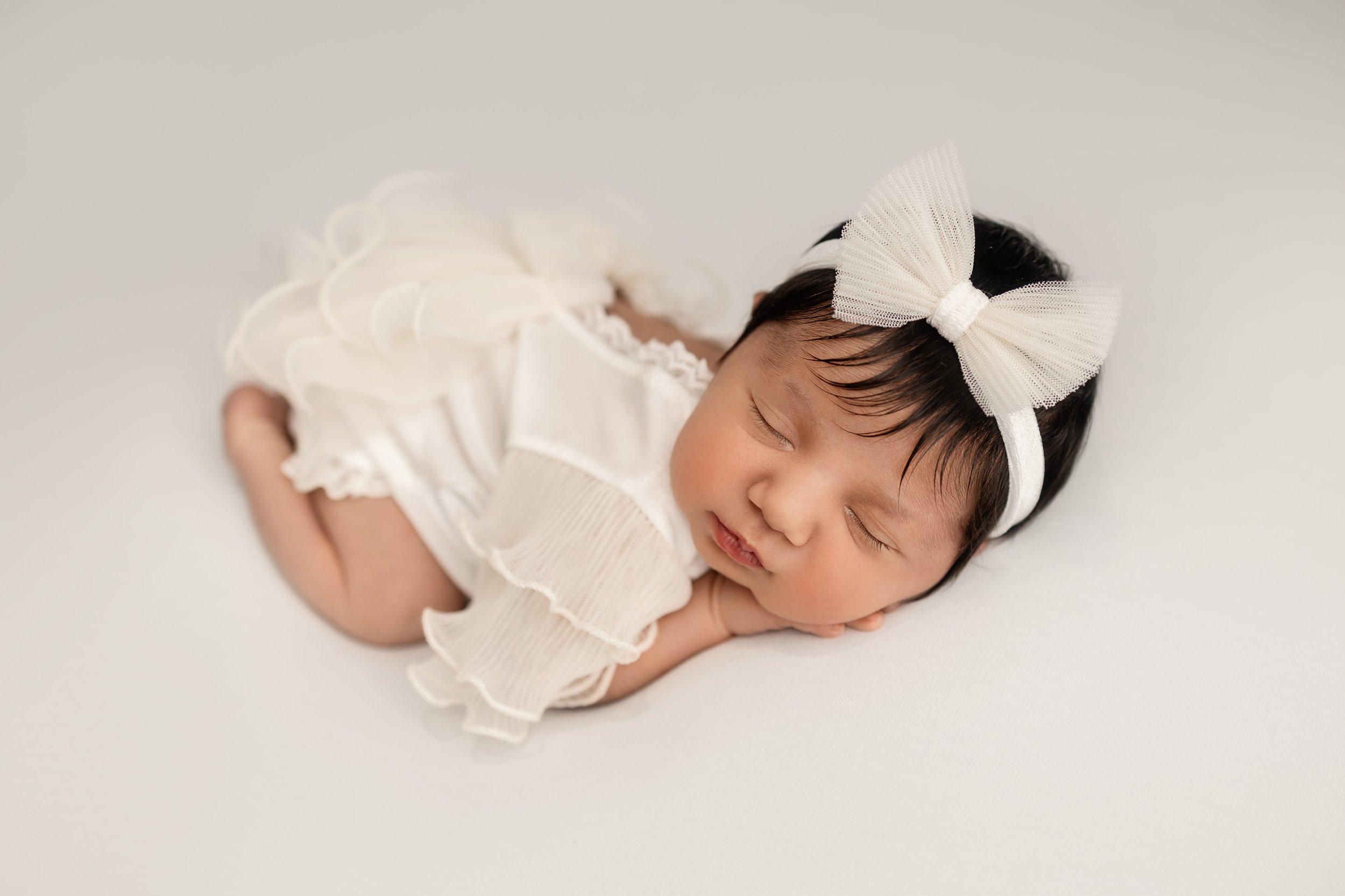 A sleeping baby girl with a bow headband, wearing a white ruffled dress, lying on a white surface.