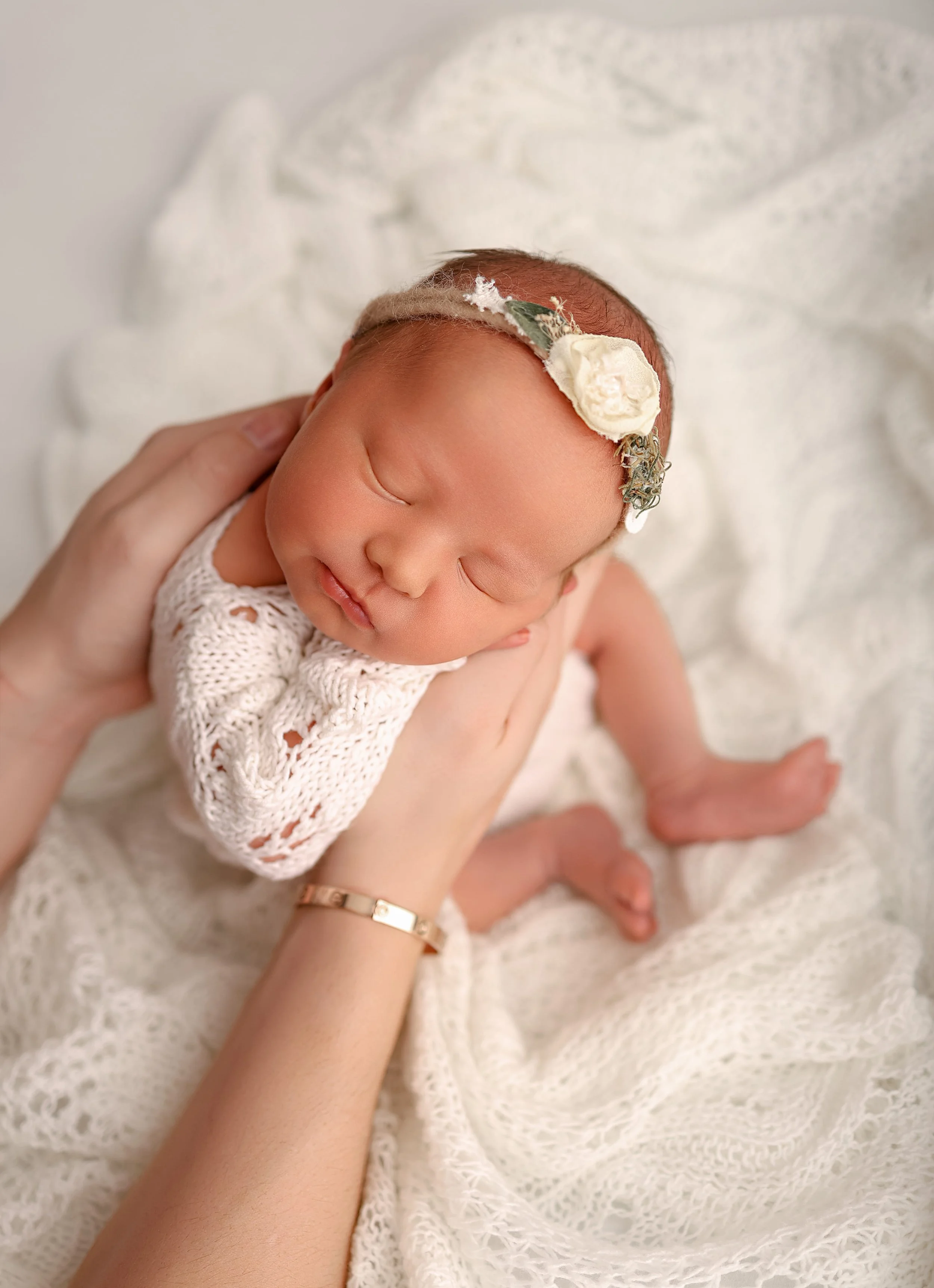Close-up of a sleeping newborn baby with a flower headband, cradled in a person's hands, lying on a soft white blanket.