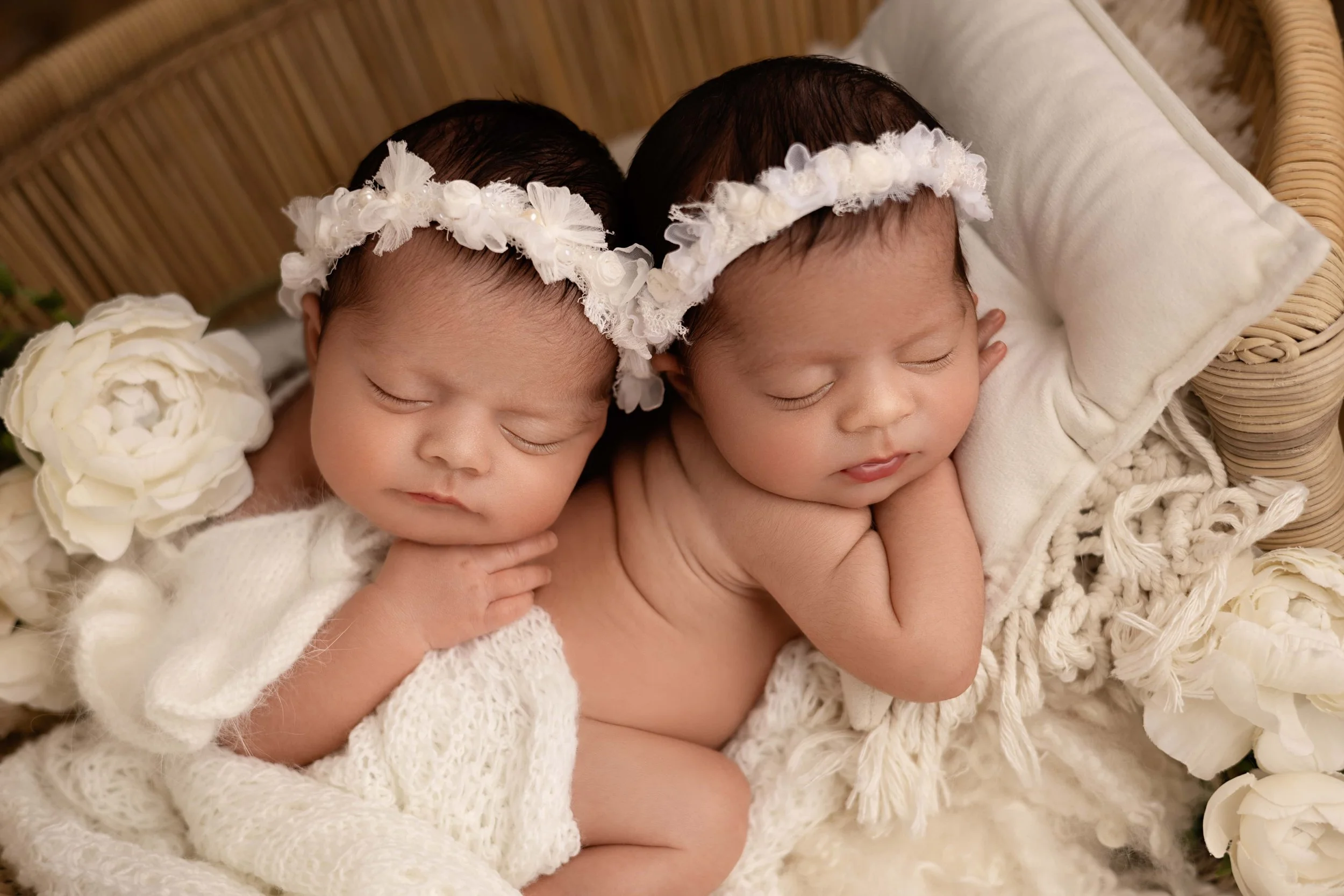 Two sleeping newborn babies with floral headbands, lying on soft blankets surrounded by white flowers.