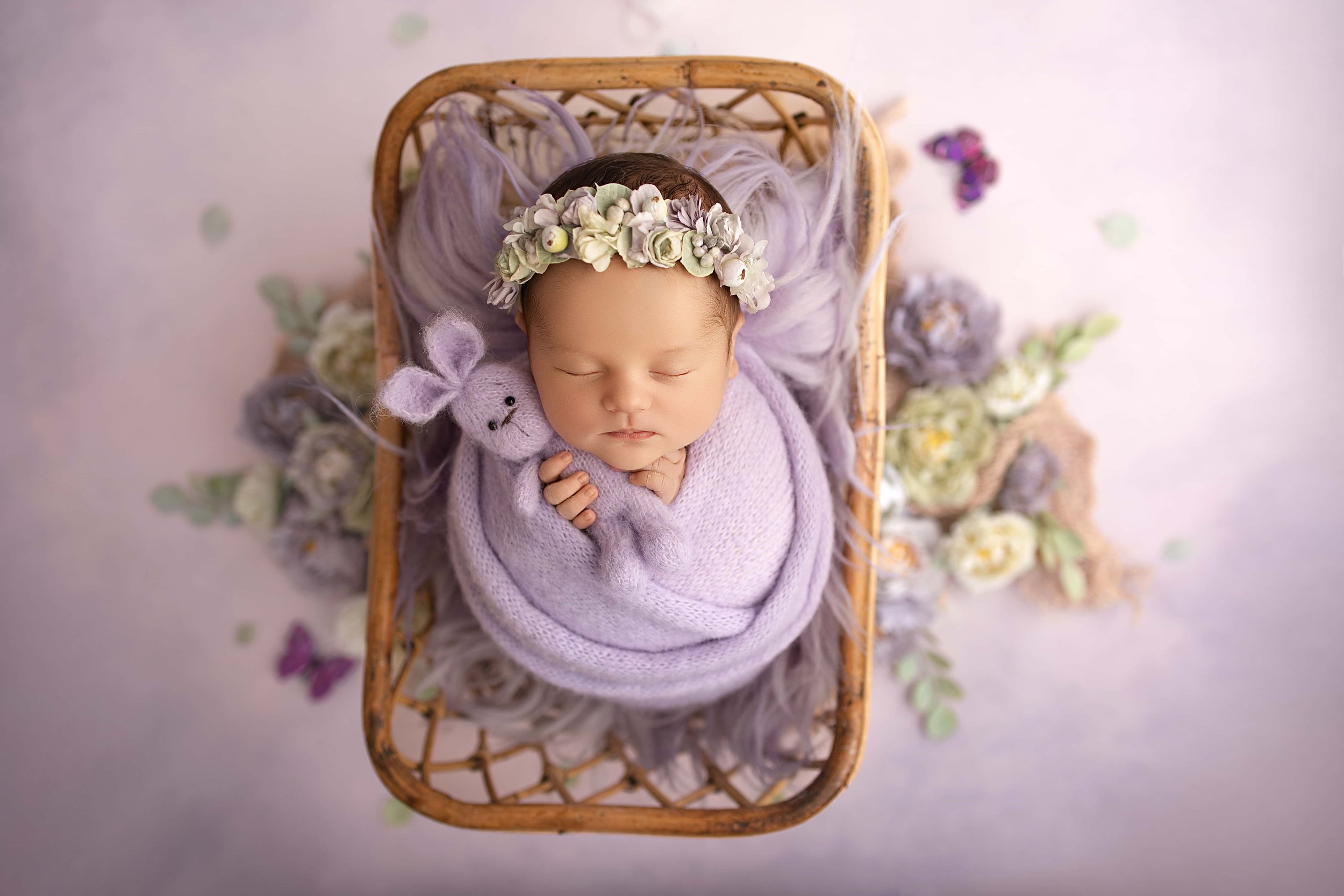 Baby dressed in a lavender wrap, with a floral headband, lying in a wicker basket surrounded by flowers and soft fabric, holding a small lavender stuffed animal with bunny ears.