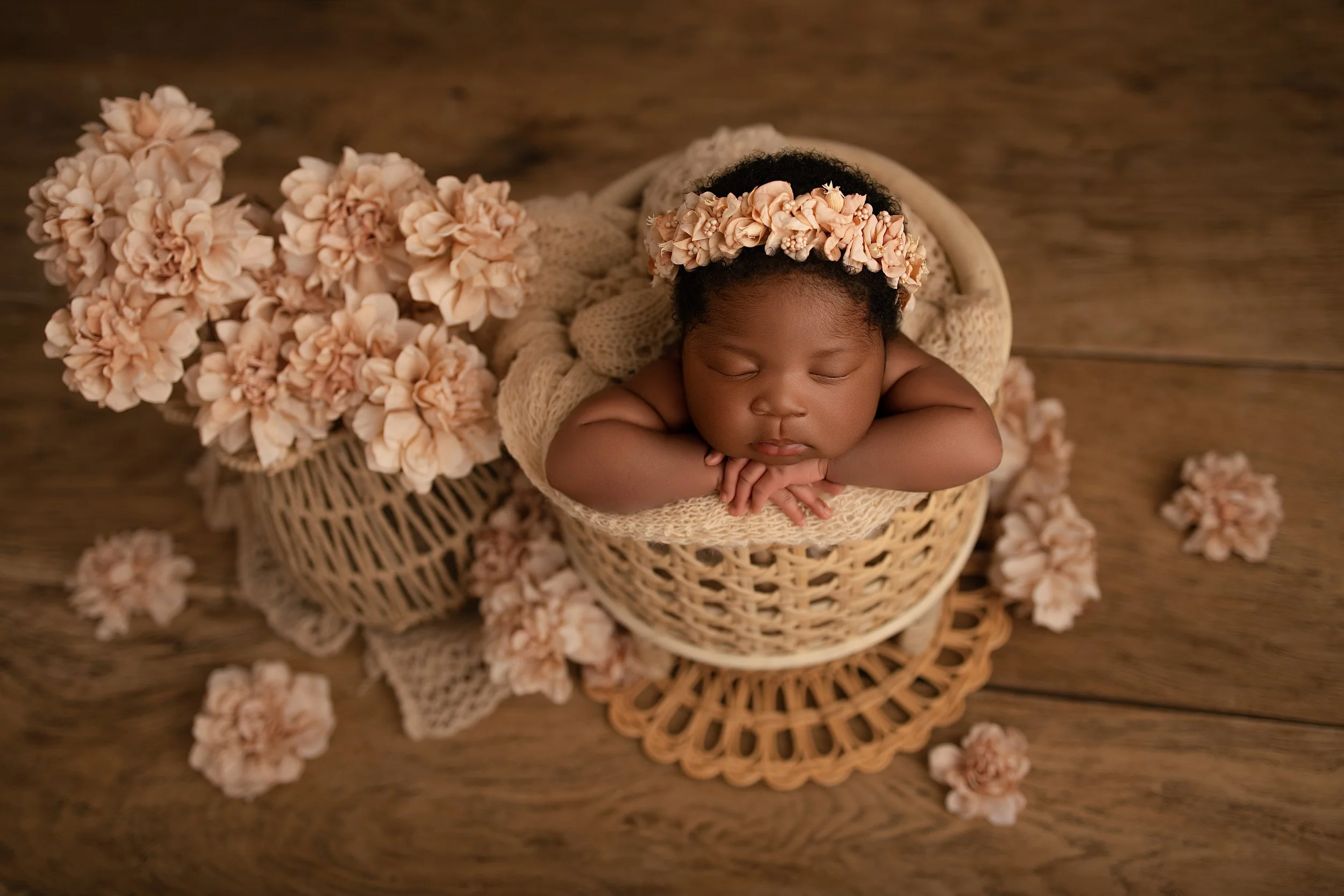 A sleeping baby girl with a flower headband, resting her chin on her folded arms inside a wicker basket with flowers and lace around her, on a wooden floor.