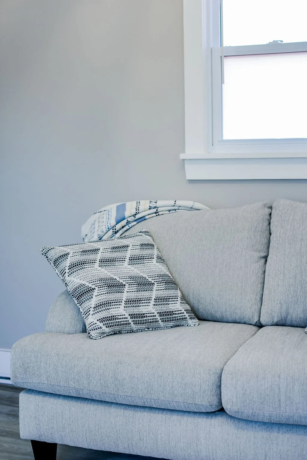 A light gray sofa with patterned throw pillows and a blanket, next to a window with white trim, in a minimalist living room.