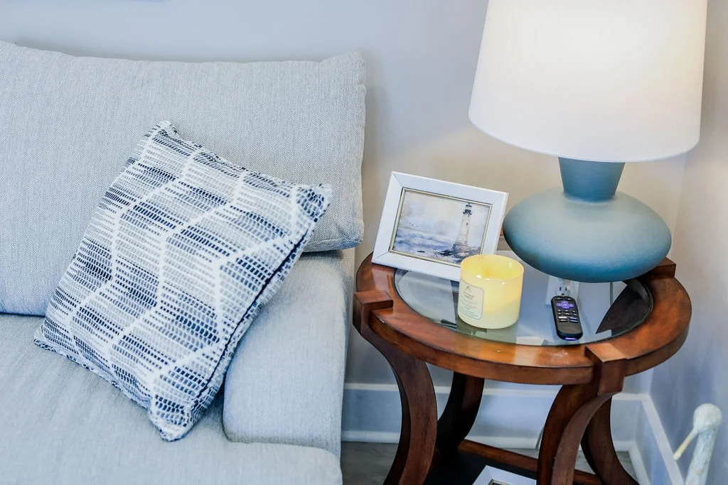 Living room corner with a grey sofa, a patterned cushion, a wooden side table with a glass top, a white lamp, a framed lighthouse photo, a yellow candle, and a TV remote.