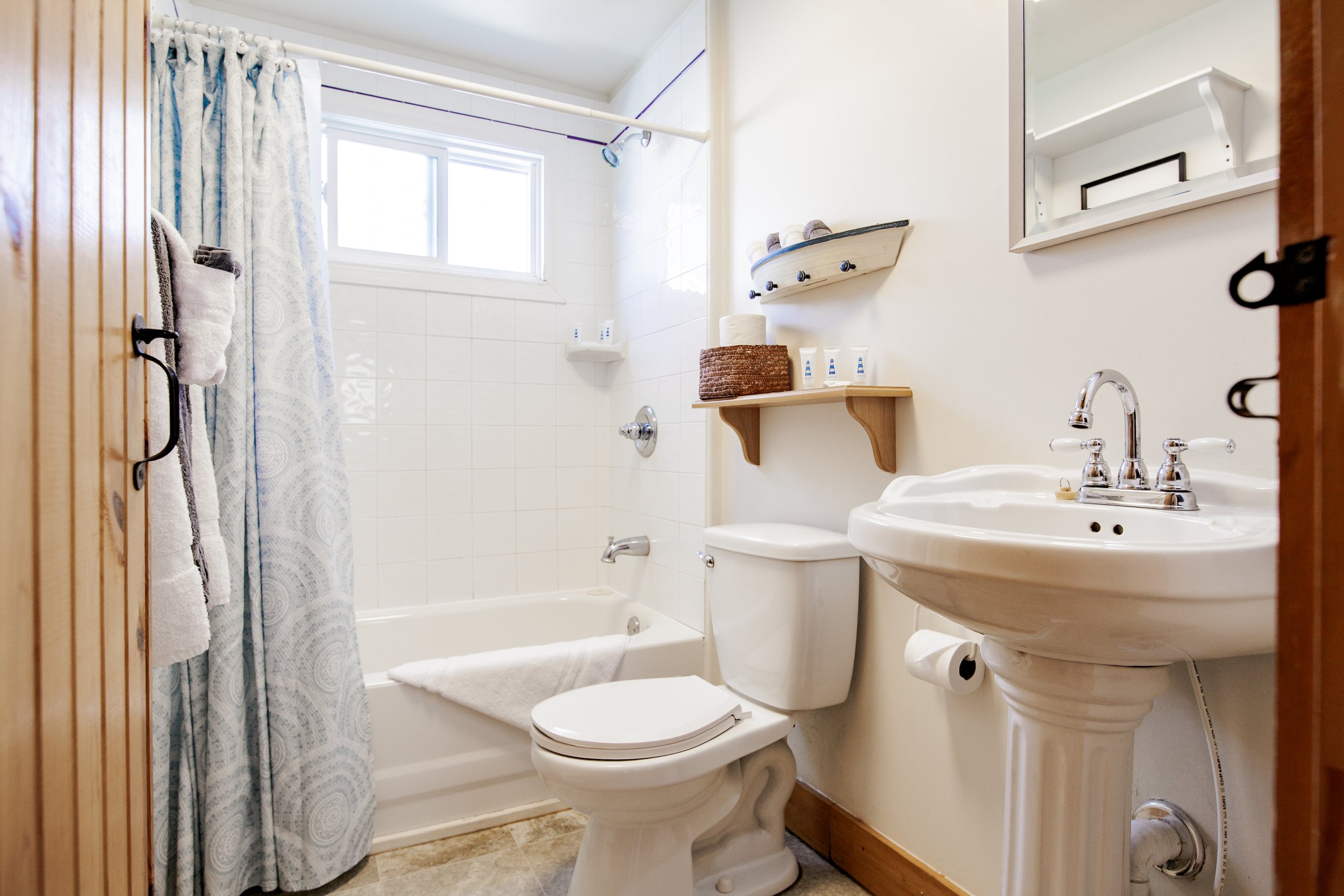 Small bathroom with a bathtub, a toilet, a pedestal sink, a mirror, a wooden shelf with toiletries, and a light-colored shower curtain, illuminated by natural light from a window.