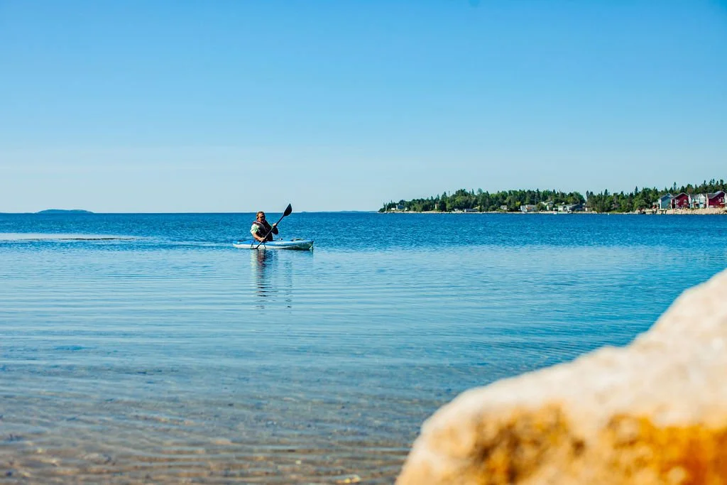 A person kayaking on calm blue water near a rocky shoreline on a sunny day, with a small town or neighborhood in the background.