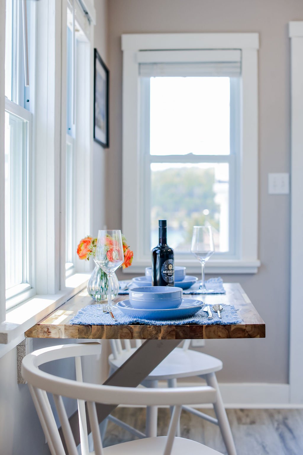 A dining table near a window with place settings, wine glasses, a bottle of wine, a small vase with flowers, and a plate and bowl arrangement, in a bright room with natural light.