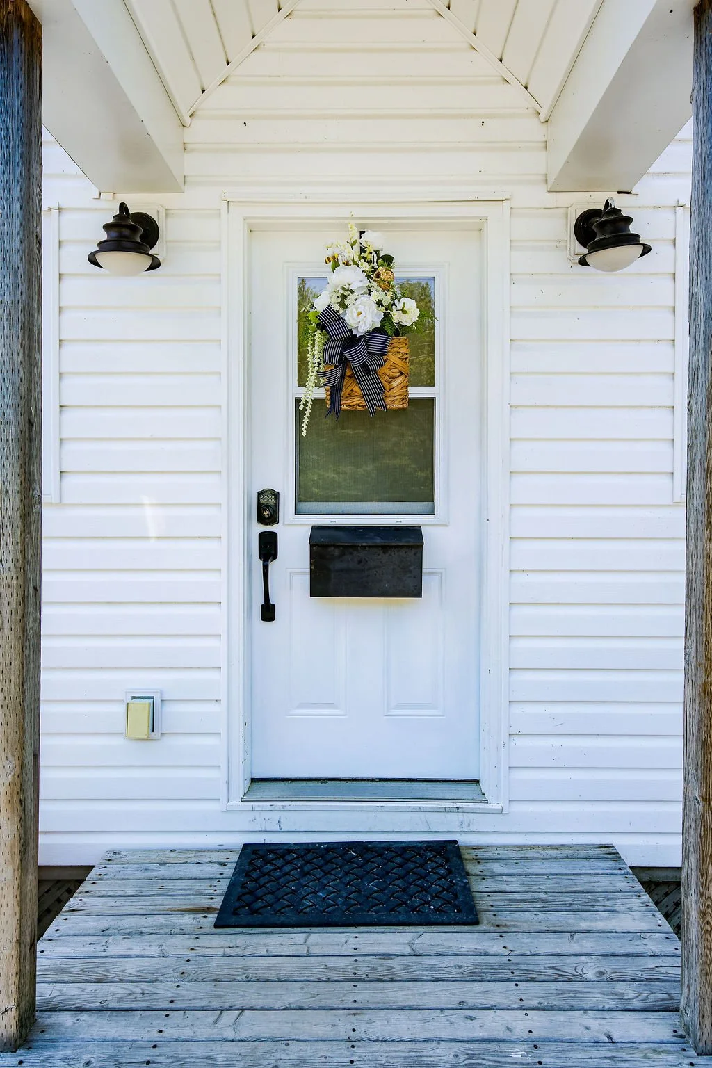 White front door with a floral wreath hanging on the window, black mailbox, black door handle, and black light fixtures on a white house with wooden steps and a black doormat.