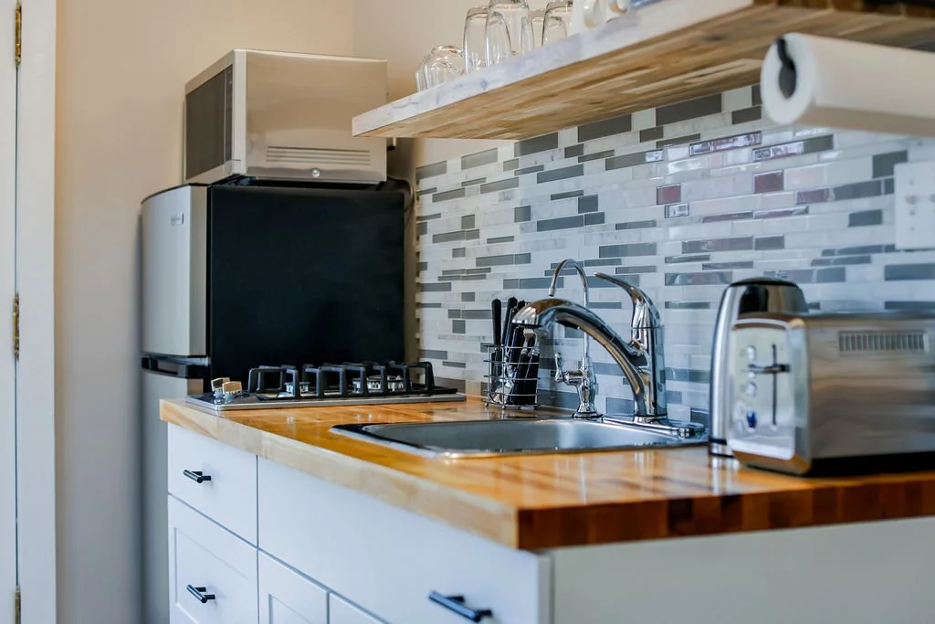 Compact kitchen with microwave, mini fridge, toaster, and cookware on a wooden countertop, backsplash with mosaic tiles, and hanging glasses