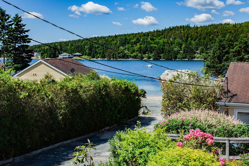 View of a lakeside neighborhood with houses, green bushes, flowering plants, a body of water, and forested hills in the background under a blue sky with scattered clouds.