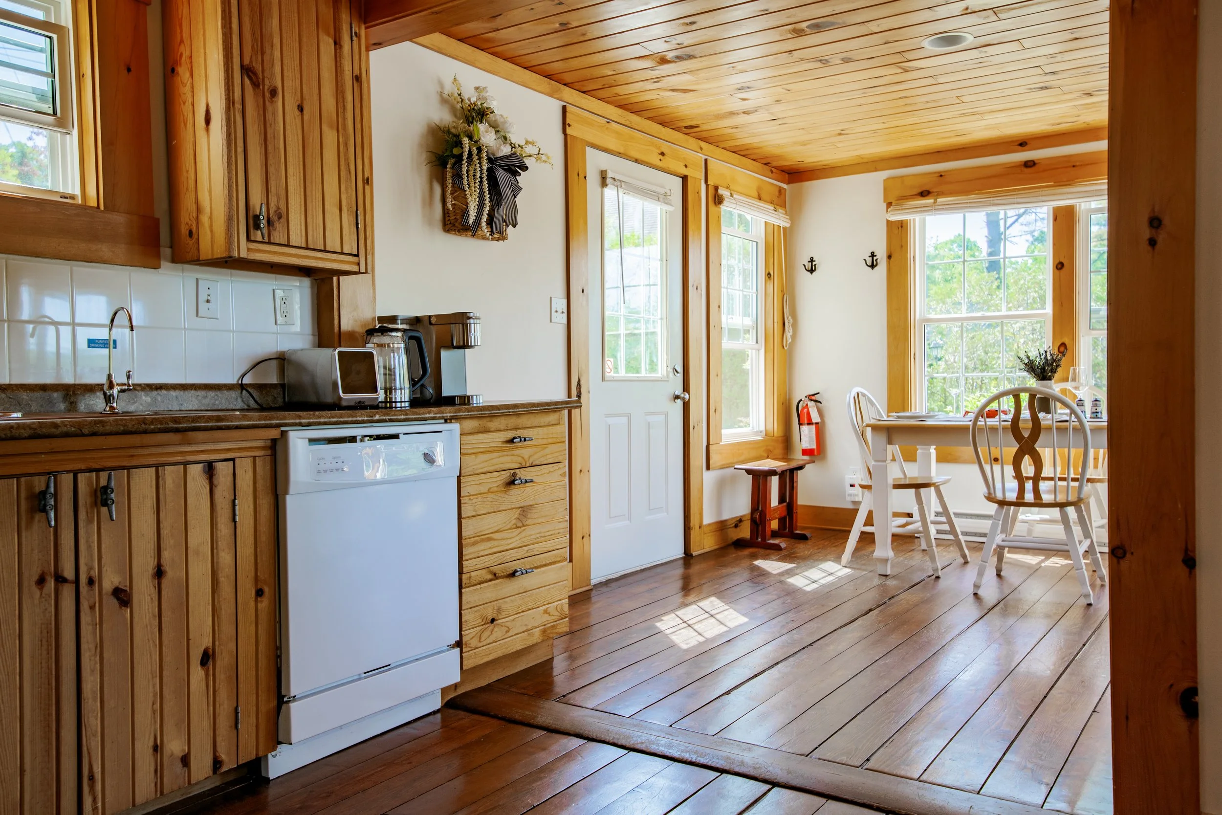 Wooden kitchen with white cabinets, white dishwasher, small kitchen appliances, and a dining area with white chairs and a table near large windows.