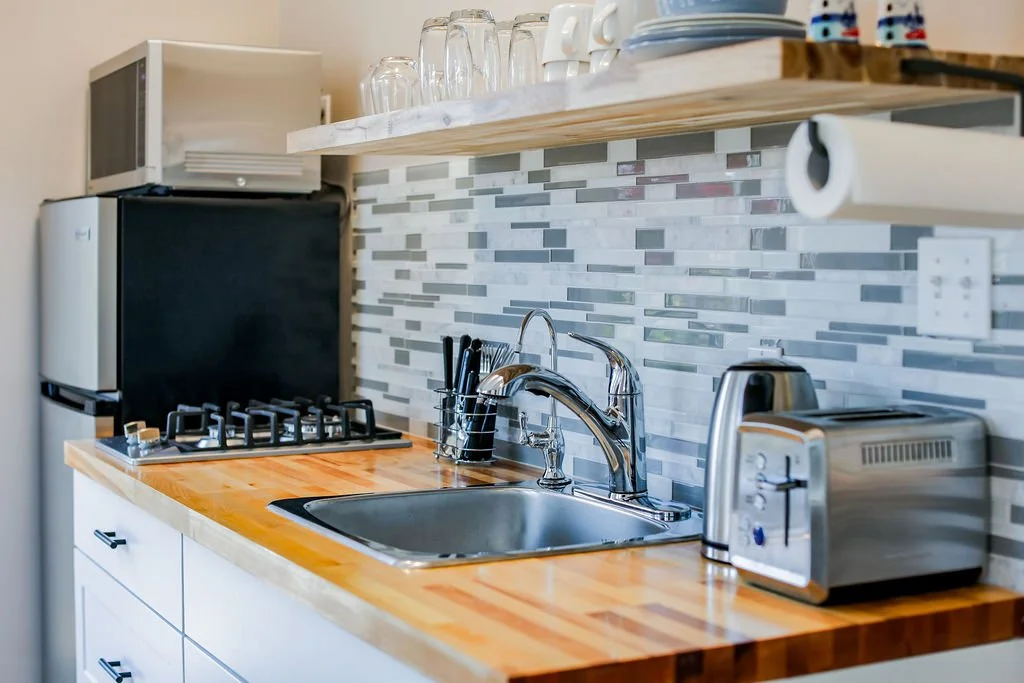 Kitchen countertop with a sink, toaster, microwave, small refrigerator, and open shelves with cups, glasses, and mugs.