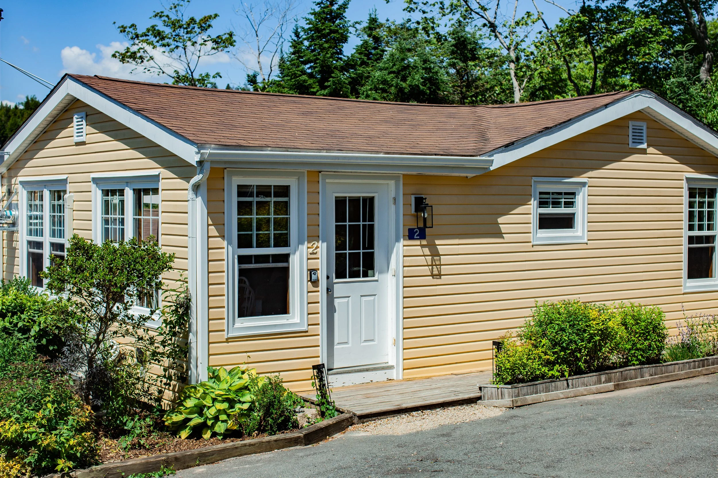 Yellow house with white trim, with a small wooden porch in front, surrounded by green shrubs and plants, and blue sky overhead.