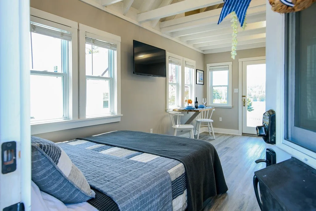 A cozy bedroom with a bed, multiple windows, a TV on the wall, and a small table with chairs near the window.