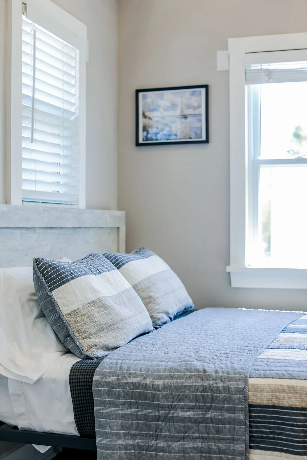 A neatly made bed with gray and white striped pillows and a gray quilt in a bright bedroom. There are white window blinds and a framed landscape painting on the wall.