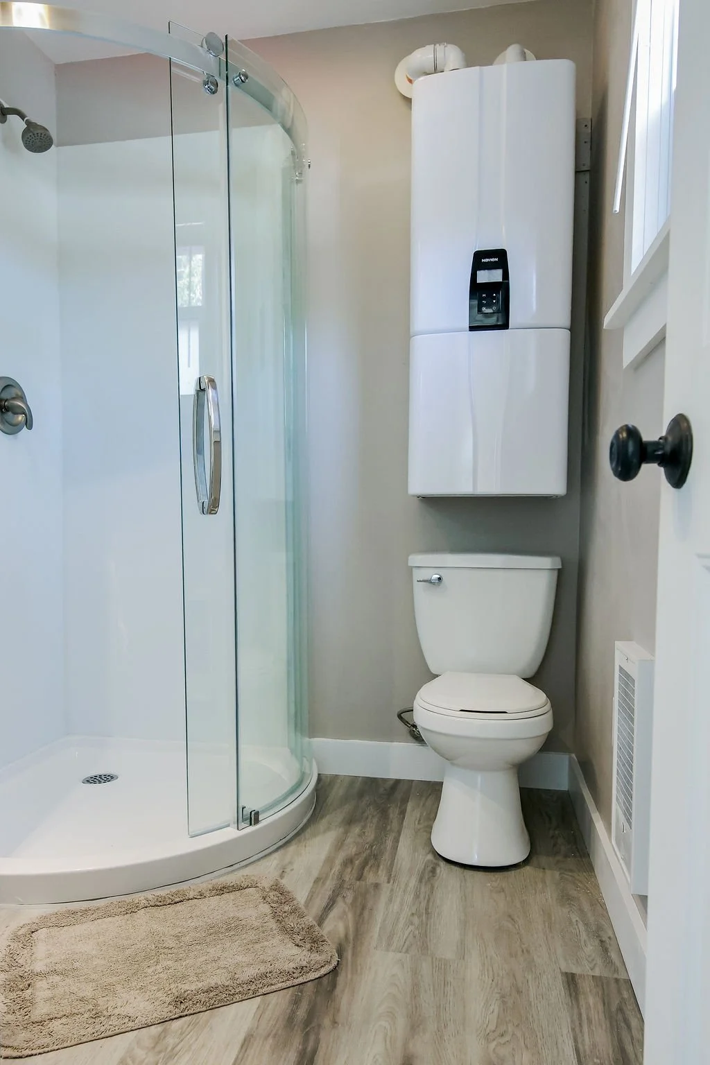 Bathroom with a shower stall, toilet, and wall-mounted water heater, with a beige bath mat on wooden flooring.