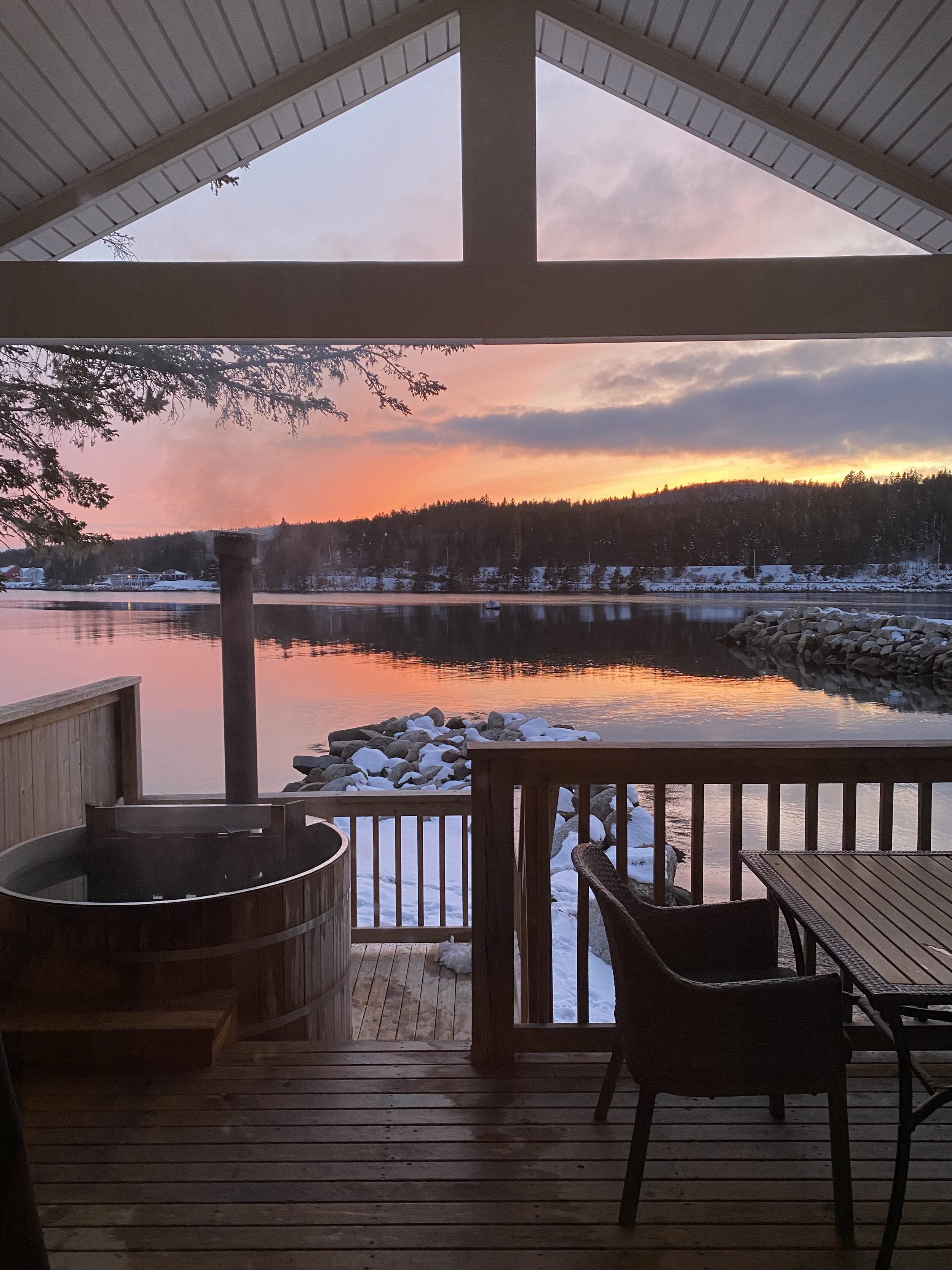 View from a wooden deck showing a hot tub, a table with chairs, snowy rocks, and a body of water with a sunset sky in the background.