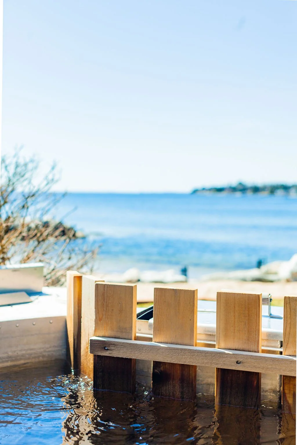 A wooden hot tub filled with water sits on a deck overlooking the ocean, with a distant shoreline and clear blue sky in the background.