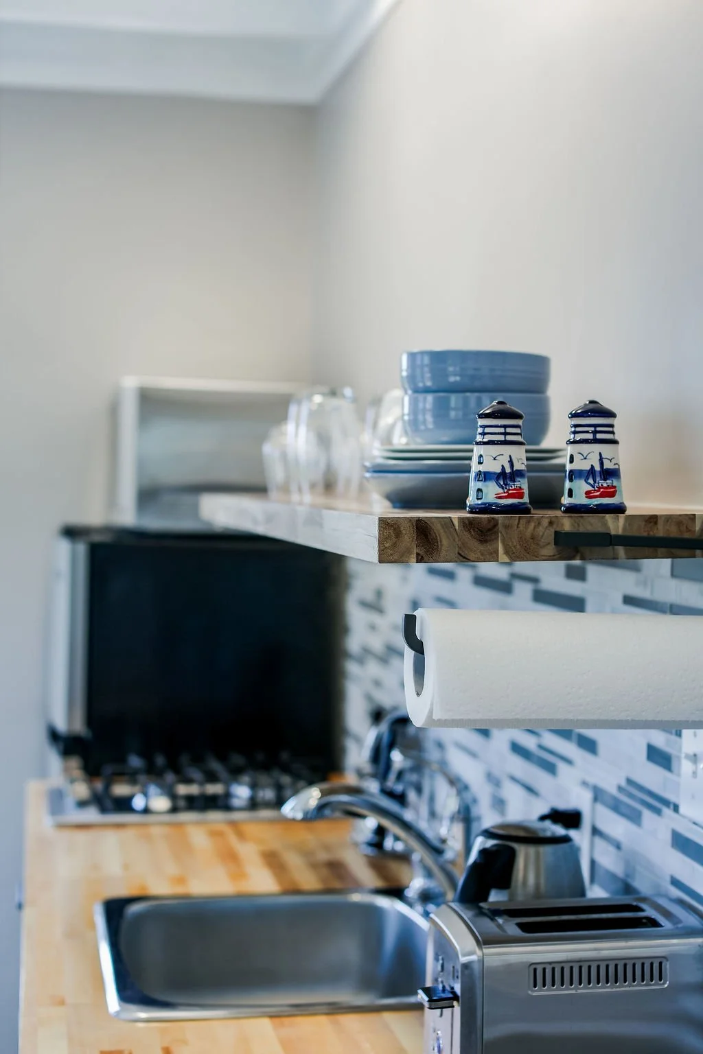 Close-up of a wooden kitchen counter with a stainless steel sink, a toaster, a paper towel roll, blue and white dishes, and decorative lighthouse salt and pepper shakers, with a stove and backsplash in the background.