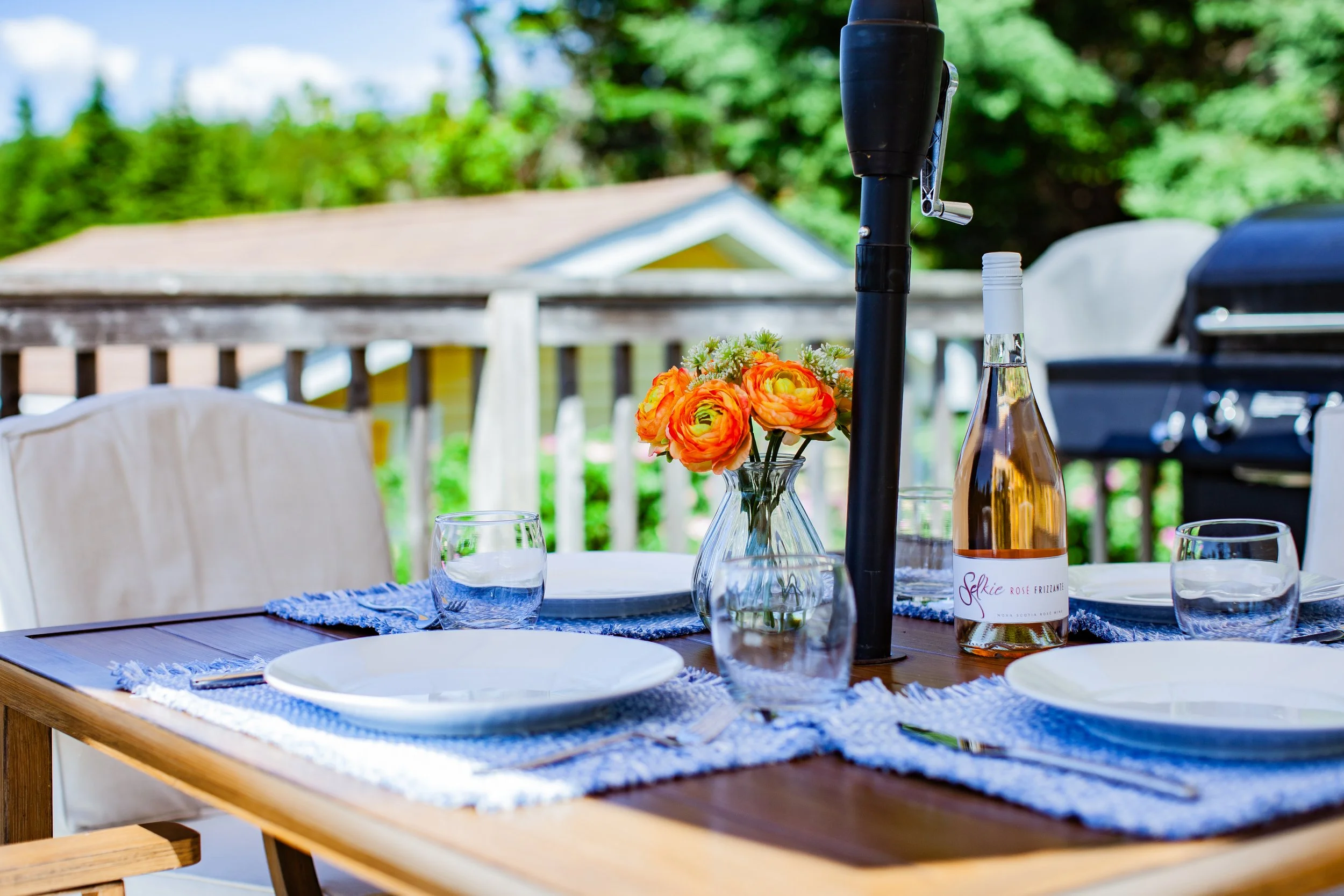An outdoor dining table set with white plates, drinking glasses, a vase with orange flowers, a bottle of rose wine, and a black patio heater, on a wooden deck with a railing and trees in the background.