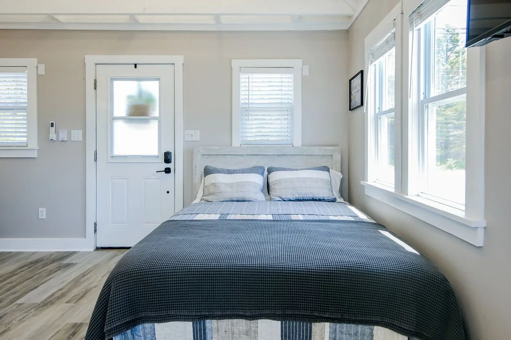 A bedroom with a white door, two windows on alternate sides of the bed, and a bed with striped bedding and pillows.