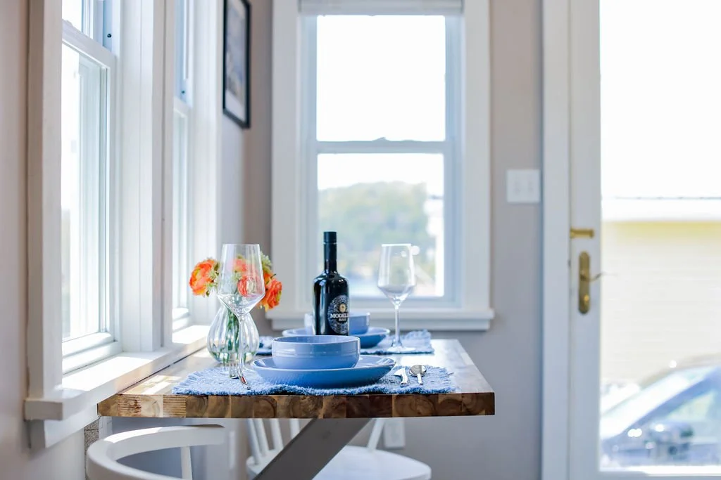 A dining table set for two with a bottle of wine, two empty wine glasses, and a small bouquet of orange and pink flowers in a glass vase, placed near a window in a bright room.