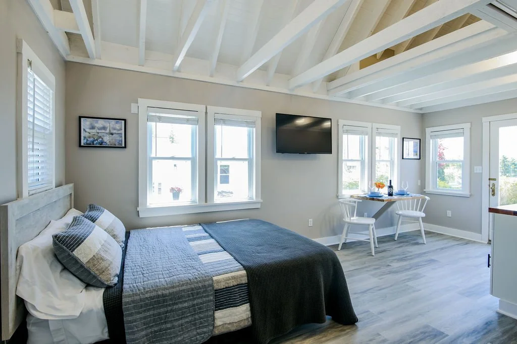 Bright bedroom with a bed, gray bedding, multiple windows, a wall-mounted television, a small dining area with two white chairs and a table, and light wood flooring.