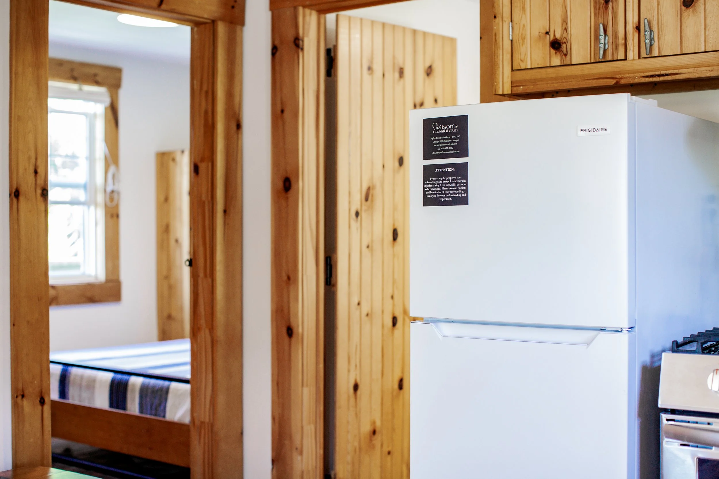 Interior of a wooden cabin with a white refrigerator, a bedroom with a bed and striped blanket, and wooden cabinets and doorframes.