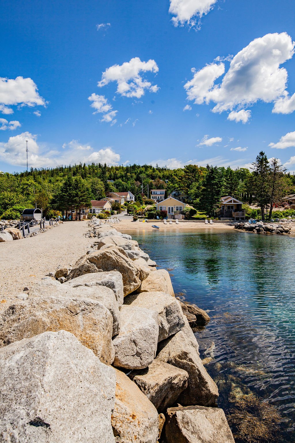 A peaceful lakeside scene with a small boat on calm water, surrounded by a rocky shoreline, green trees, and houses under a blue sky with scattered white clouds.