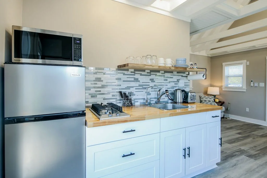 Small kitchen with a refrigerator, microwave, toaster, kettle, and utensils. Open shelving with dishes and glasses, tile backsplash, and a countertop.